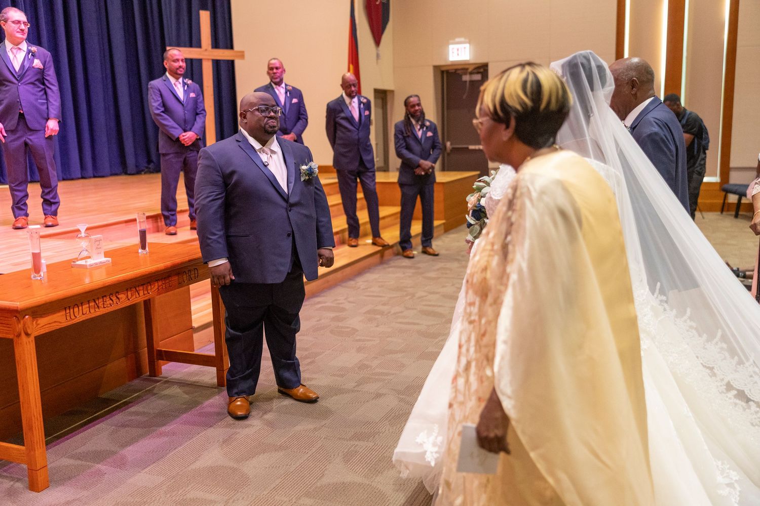 Groom stands at church altar awaiting bride in lace gown and veil, with groomsmen in purple suits on stage.