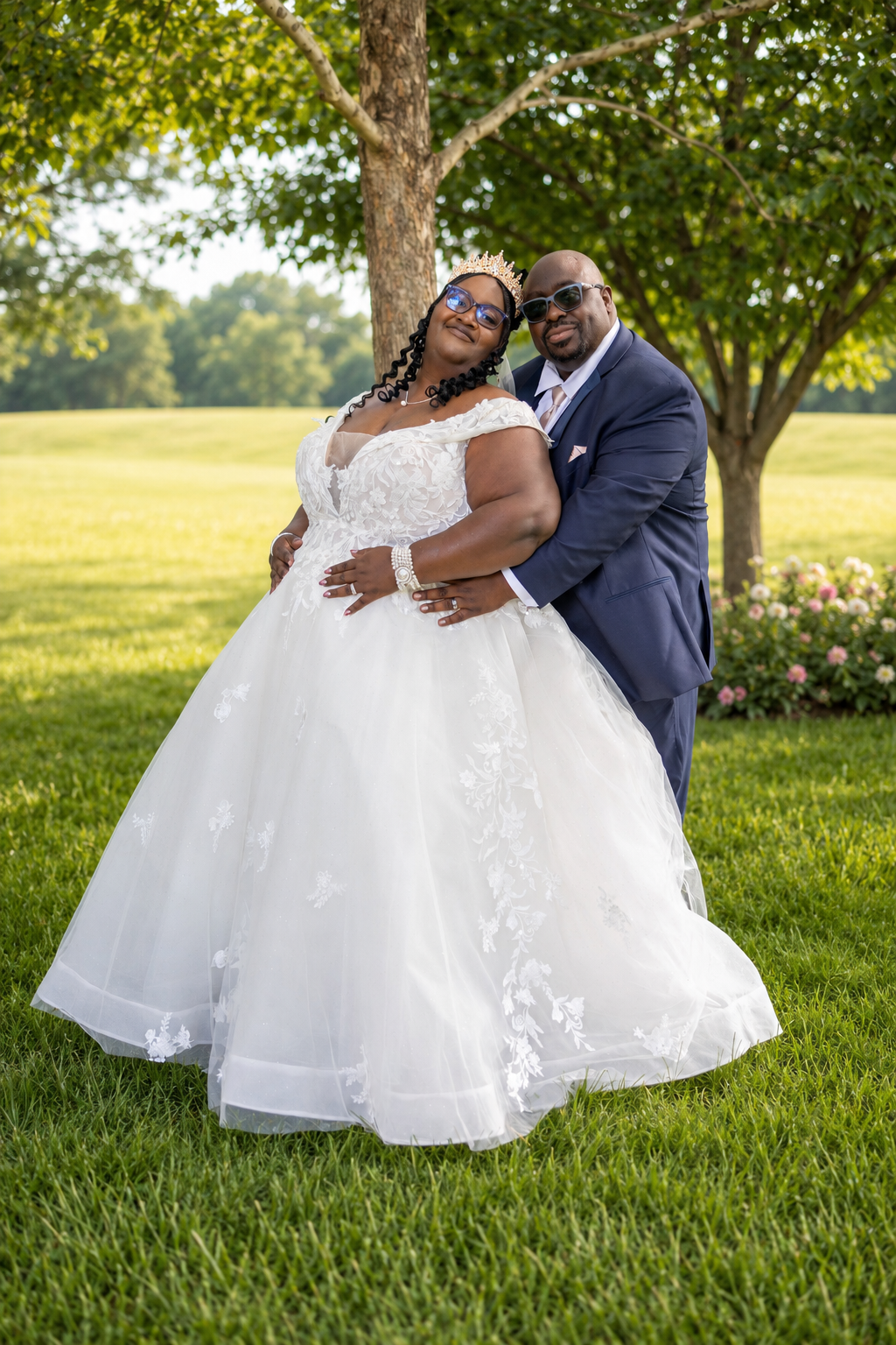 A bride in a white ball gown and groom in a navy suit pose together on lush green grass under a tree.