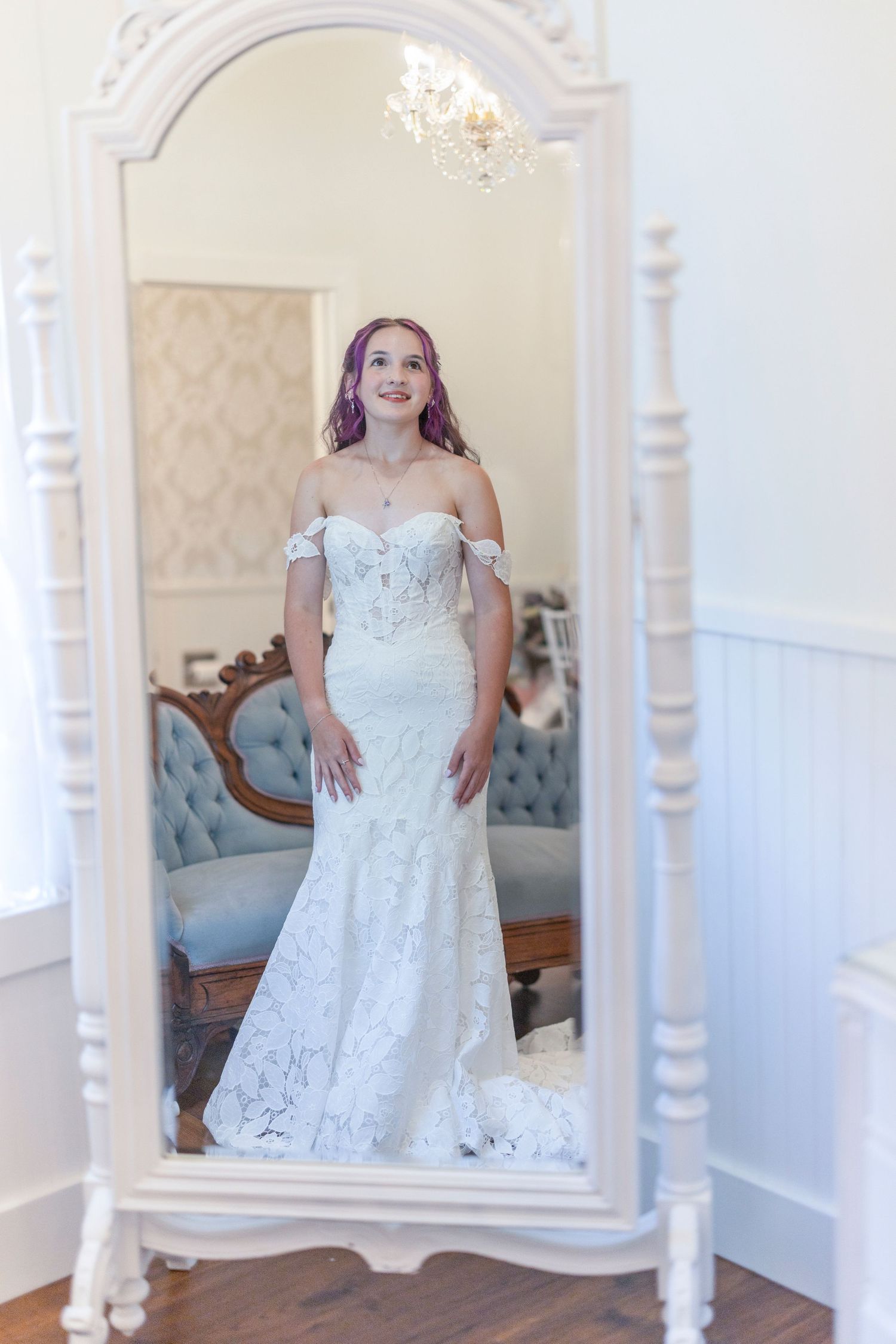 Young bride with purple hair wearing an off-shoulder lace mermaid wedding gown, posing in front of an ornate white mirror.