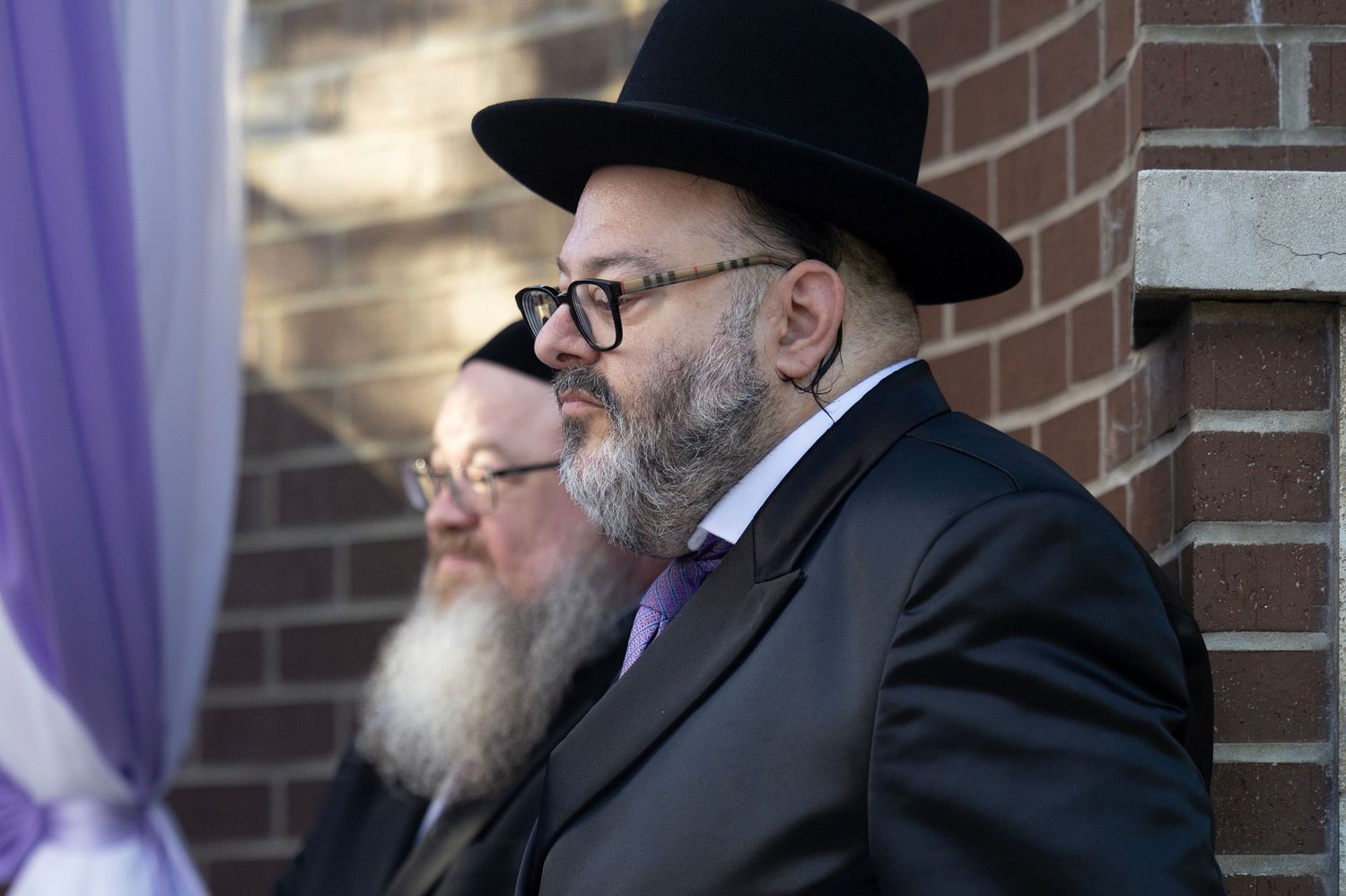 A group of Orthodox Jewish men in traditional black hats and coats gather in conversation near a brick wall.