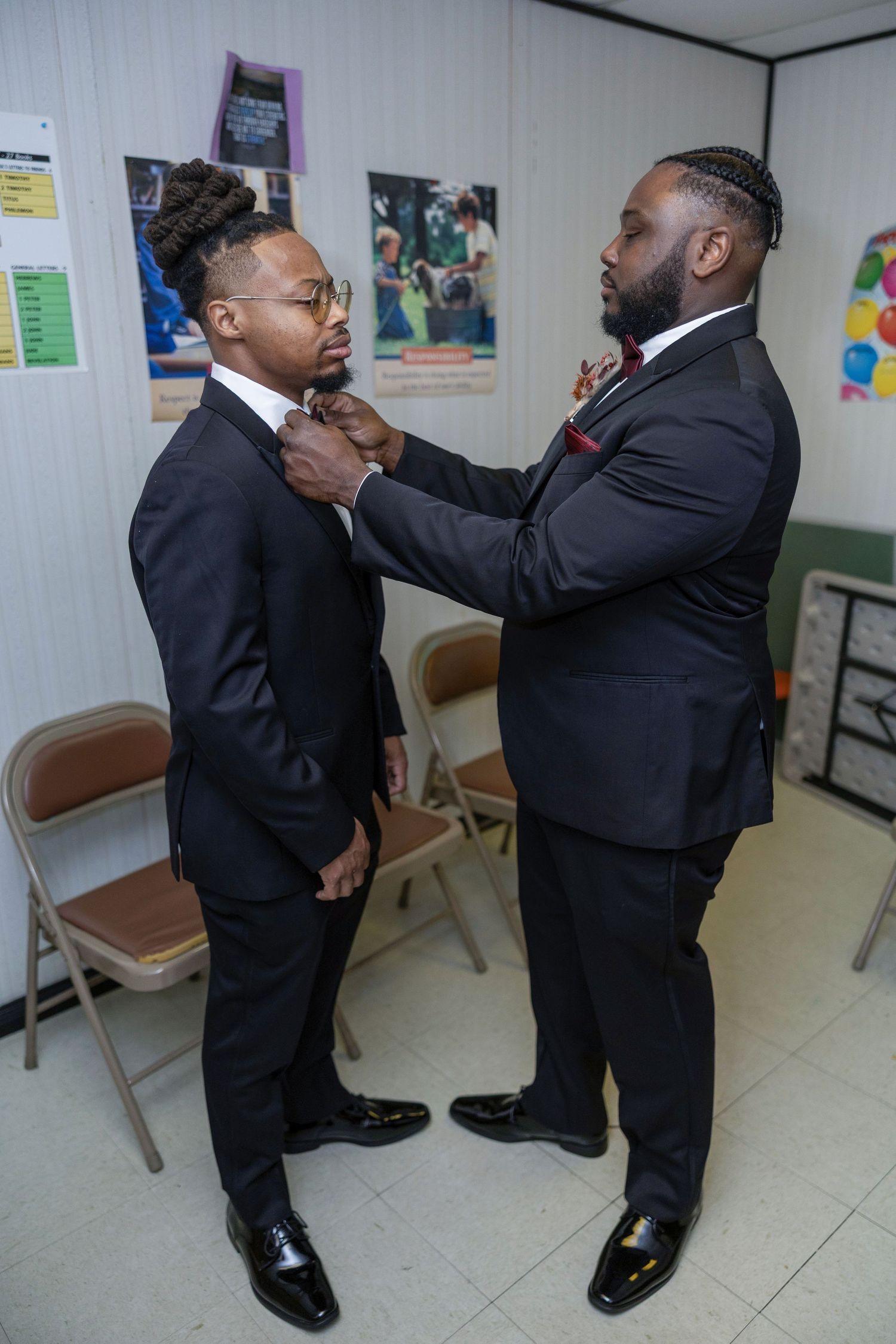 Two men in black suits practice self-defense moves in a medical office hallway with posters on the wall.