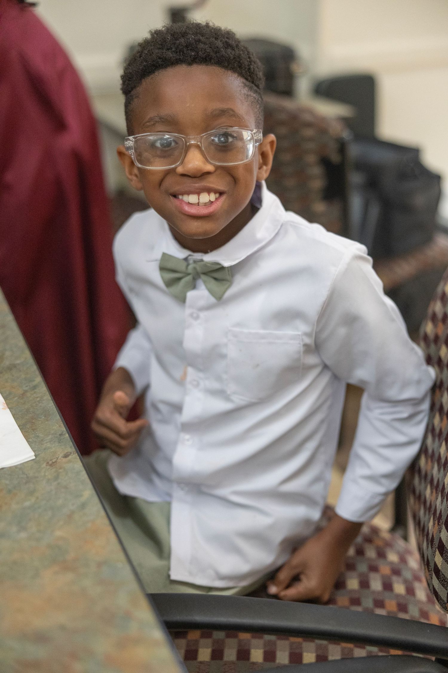 Student in white dress shirt and bow tie smiles while sitting at a desk in a classroom setting.