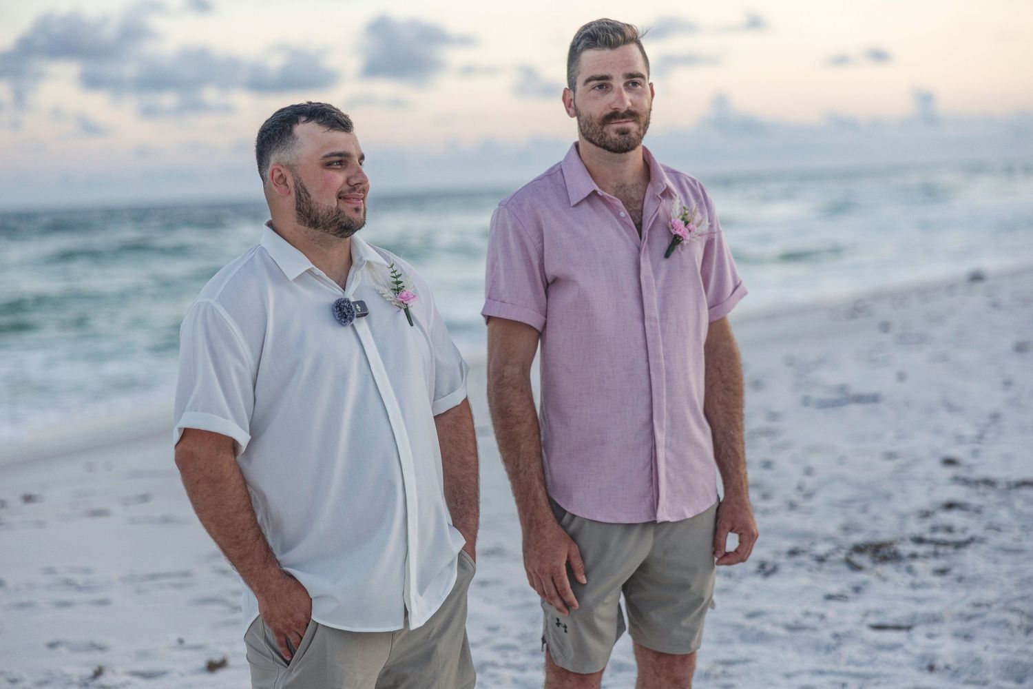 Two people walk along a sandy beach at sunset wearing light colored casual clothing and shorts.