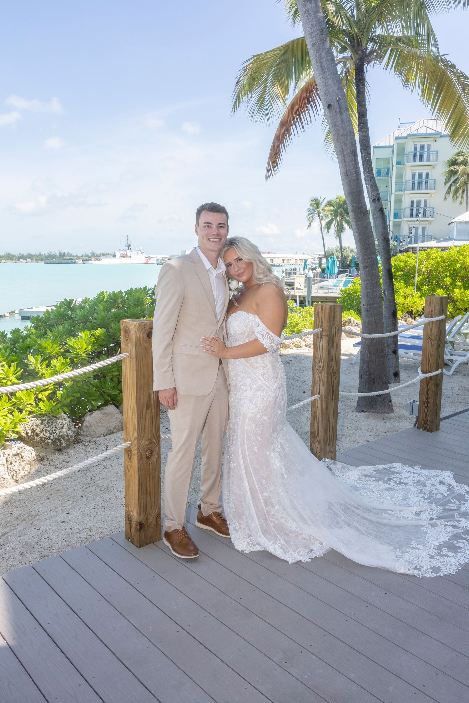 A couple poses on a wooden deck overlooking a tropical waterfront with palm trees and resort buildings in the background.