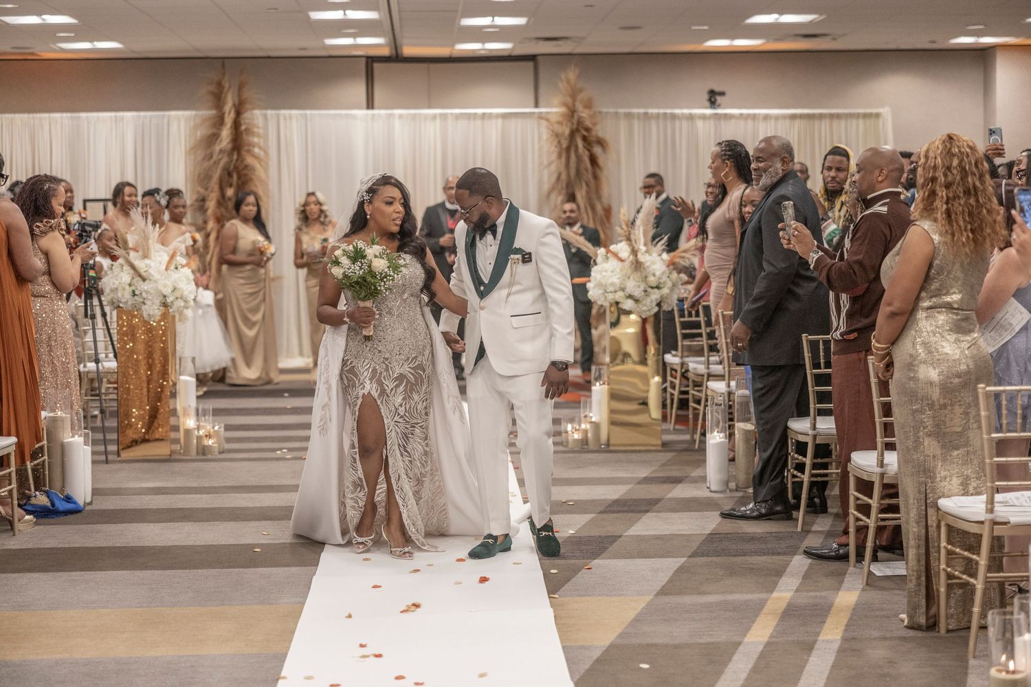 A couple walks down the aisle during their wedding ceremony while guests look on in a decorated indoor venue.