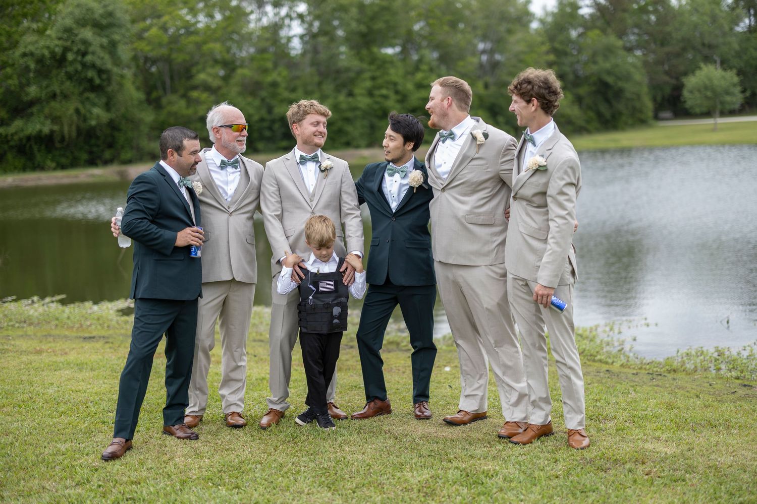 Wedding party members in light gray suits stand together on grassy lawn near a scenic pond.