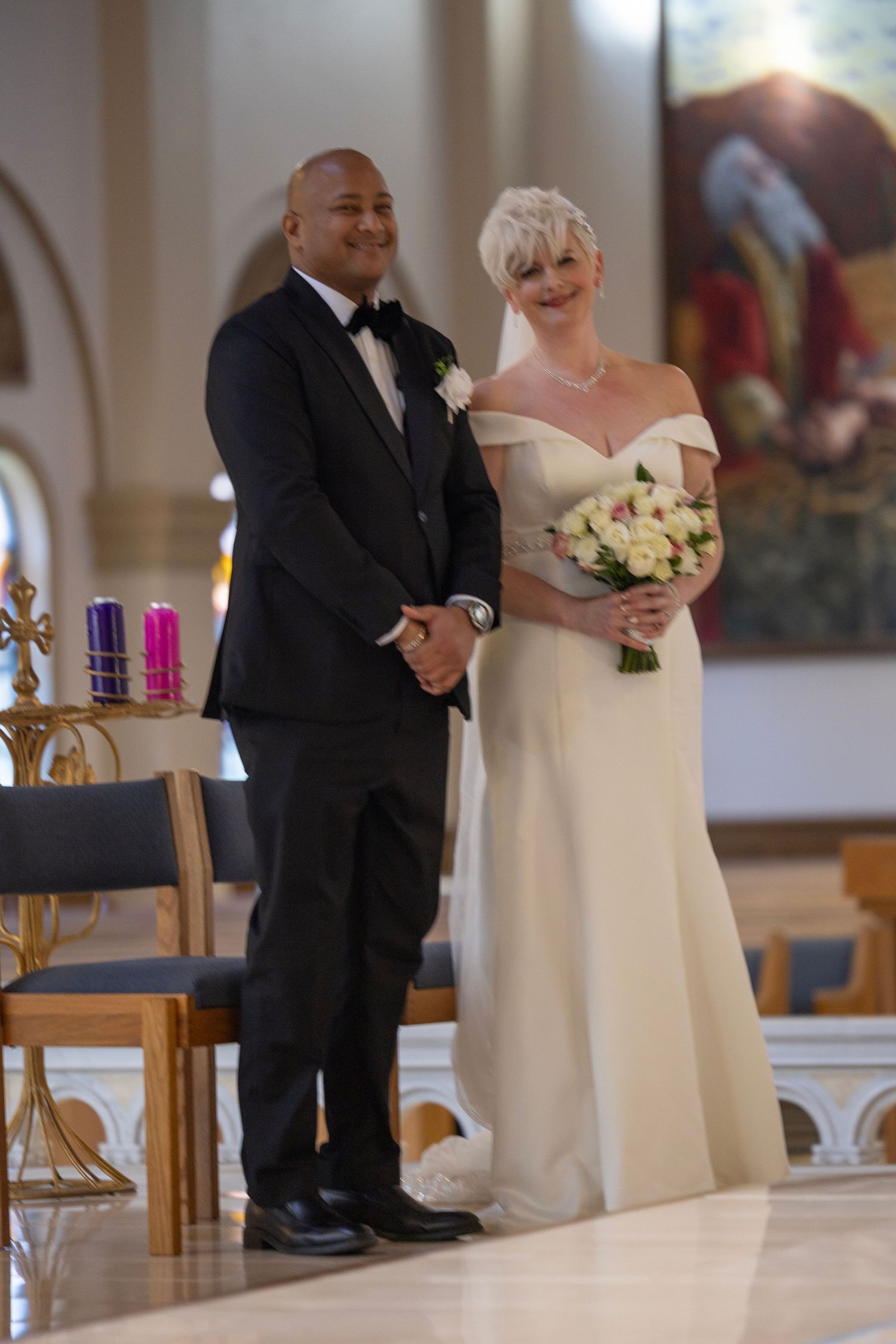 A couple stands together at their wedding ceremony, with the bride in an elegant off-shoulder gown holding a white bouquet.
