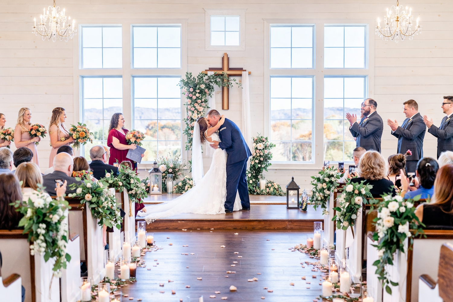 Wedding guests react with joy and emotion during a ceremony in a bright, sunlit venue with white decor and floral arrangements.