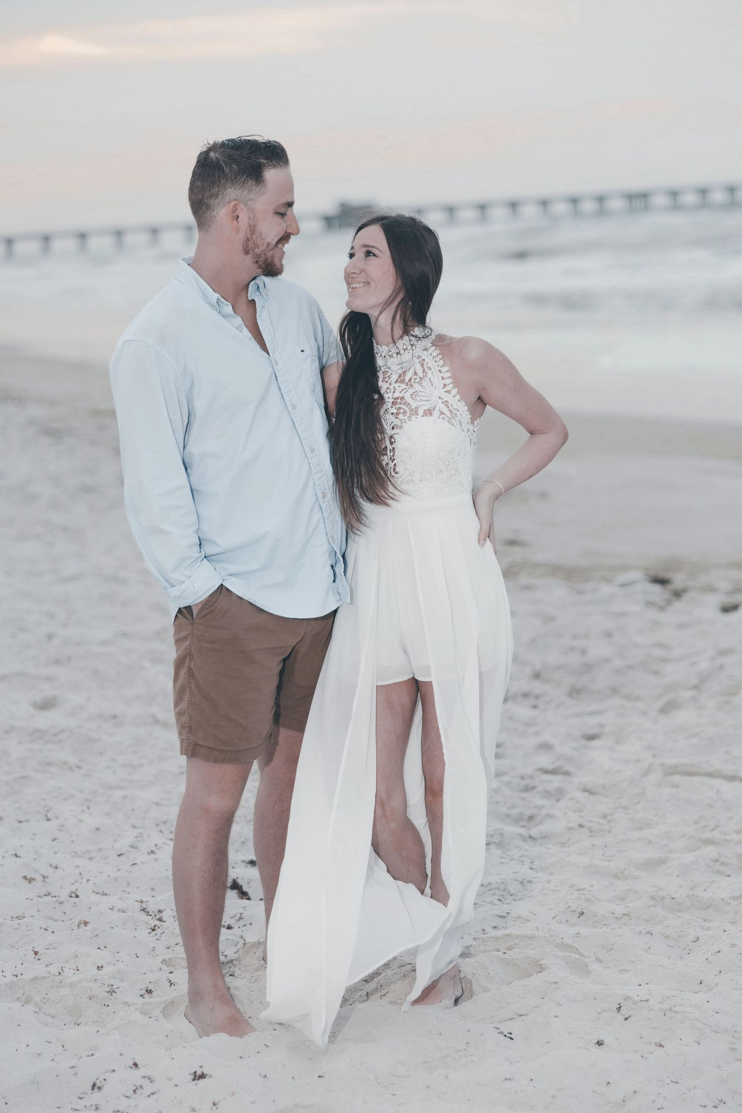 Couple in white dress and blue shirt sharing a romantic moment on a sandy beach at sunset near a pier.