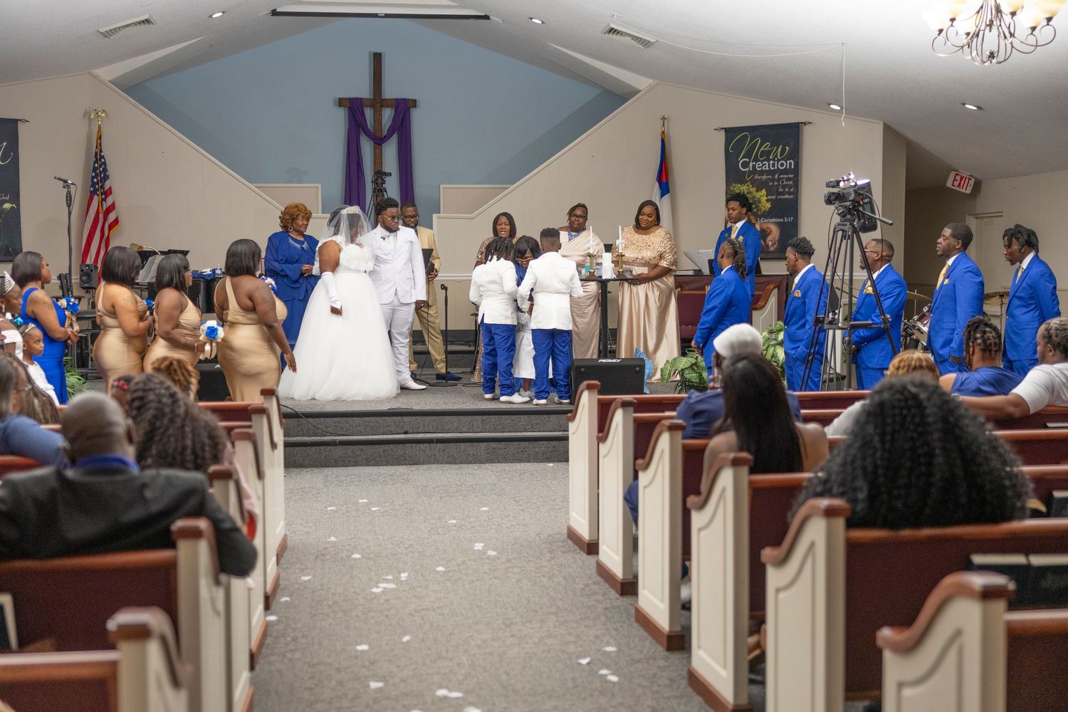 Wedding ceremony in a church with bride, groom, and wedding party in blue and gold attire before a cross.