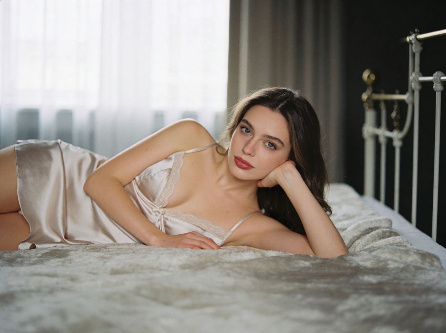 A young woman in silk lingerie poses gracefully on a bed near a window in a softly lit bedroom.