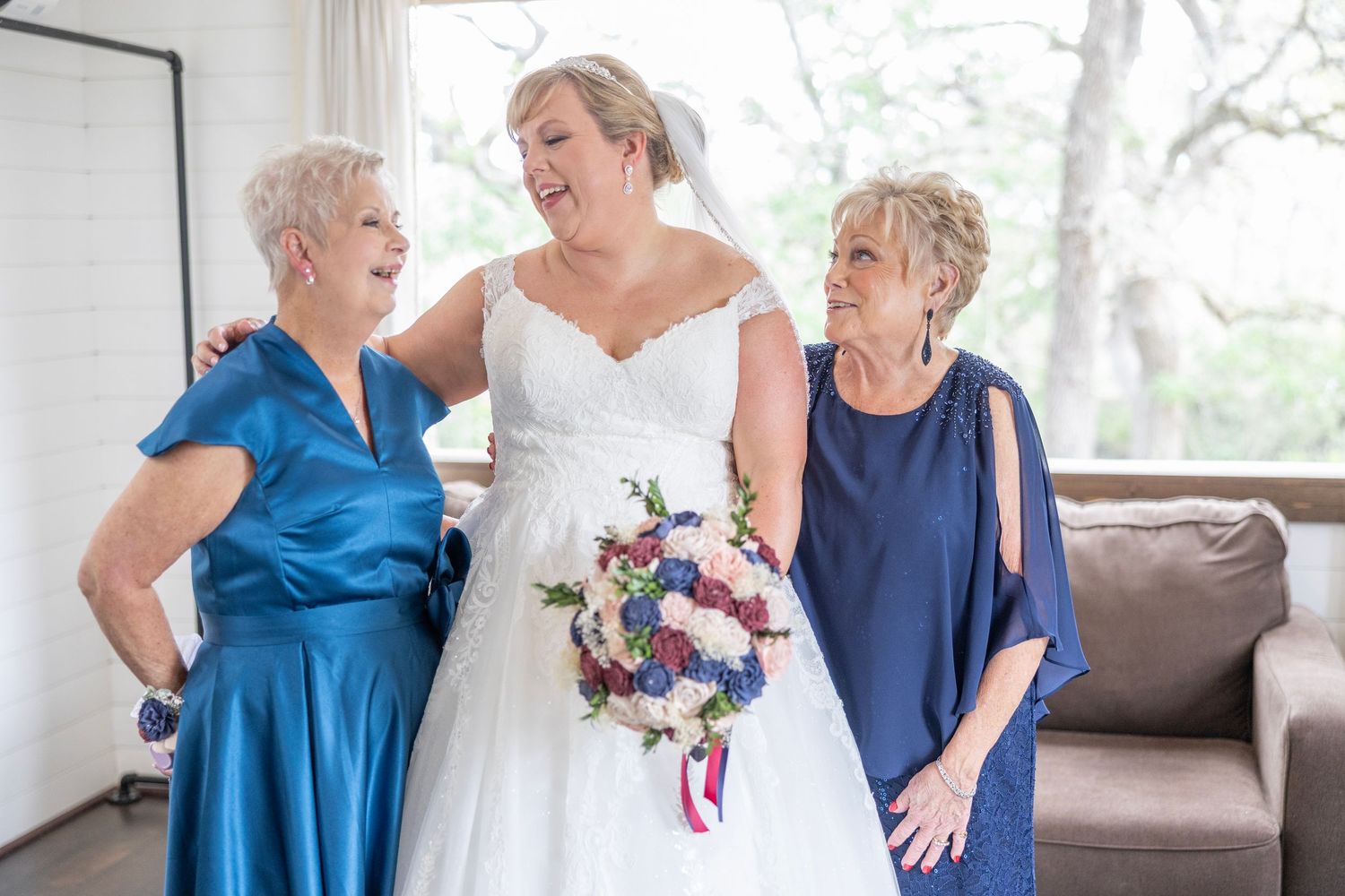 Bride in white gown holding bouquet laughs with two women in blue dresses during wedding preparations indoors.
