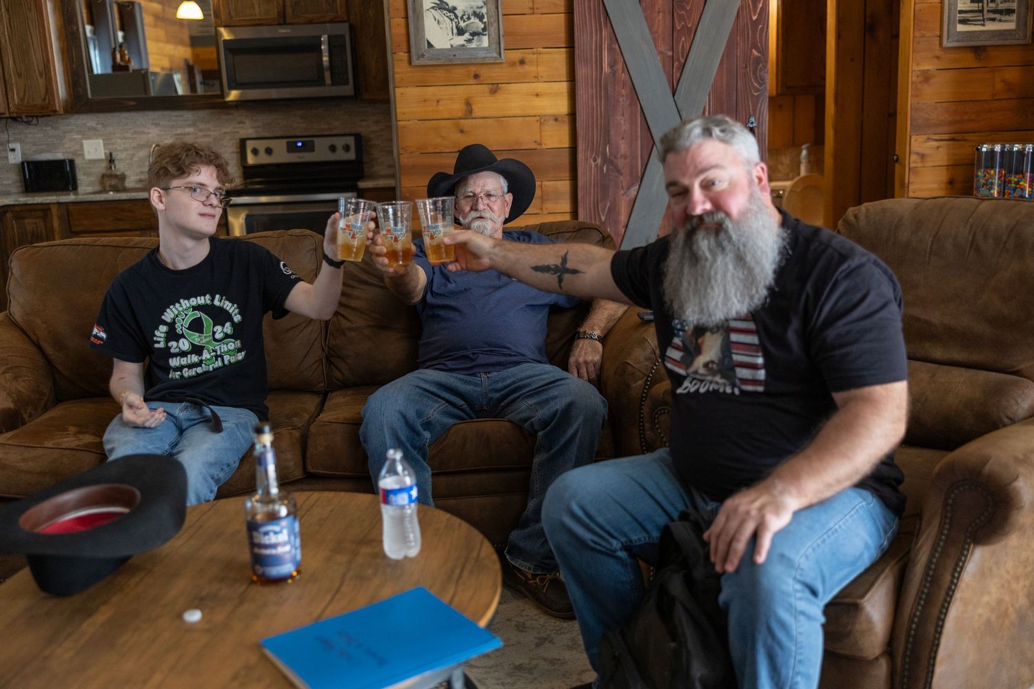 Three men relaxing on a leather couch in a rustic log cabin, raising beer glasses in a toast together.
