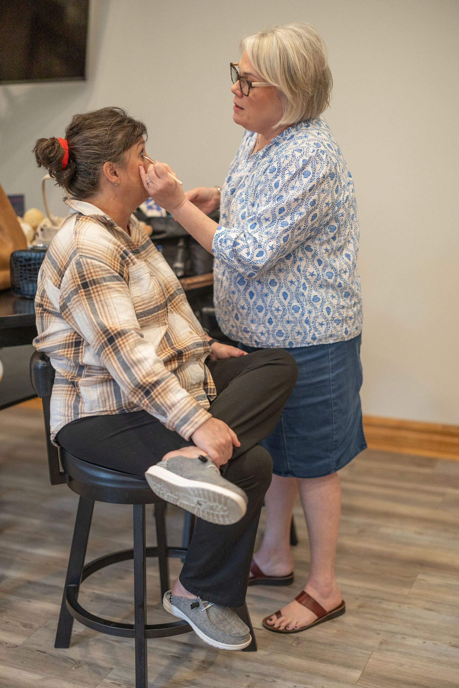 A makeup artist applies makeup to a seated woman on a stool in a salon setting.
