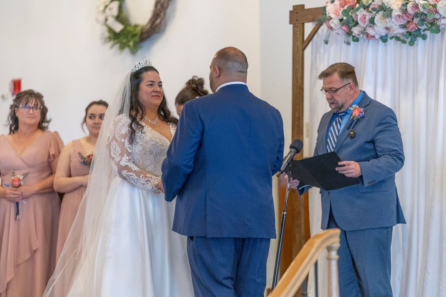 Bride and groom exchange vows at wedding altar with bridesmaids in mauve dresses and officiant holding ceremony book.