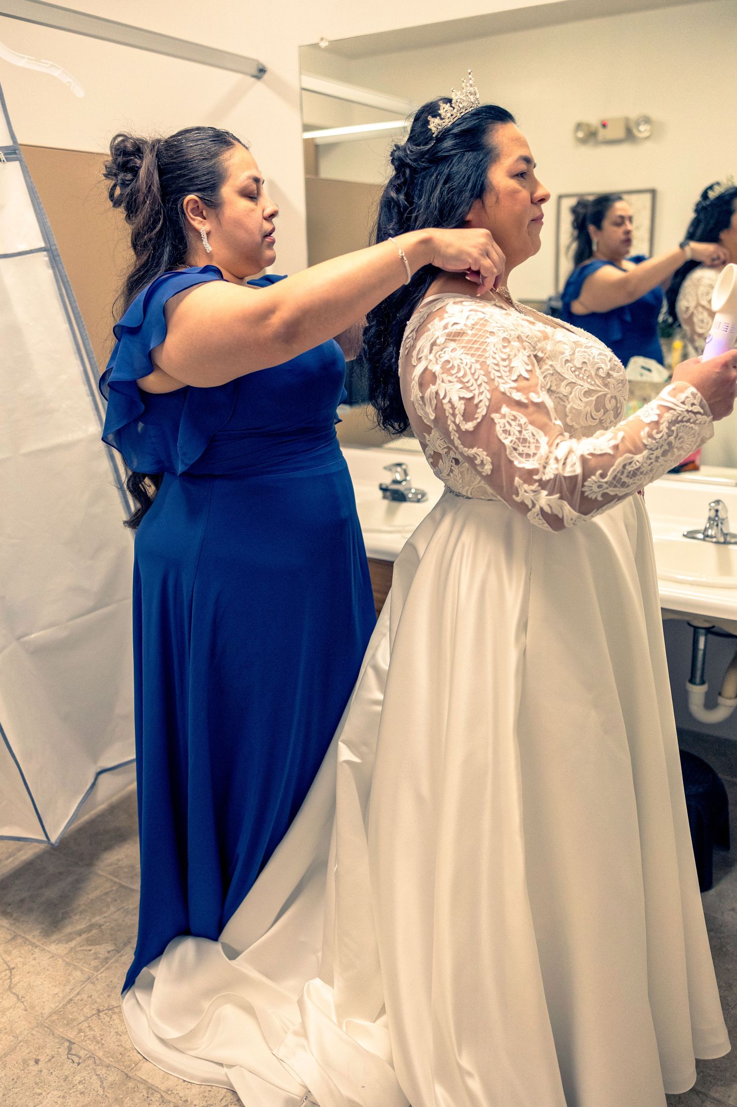 Bridesmaid in blue gown helping bride in lace wedding dress with tiara prepare in bathroom mirror.