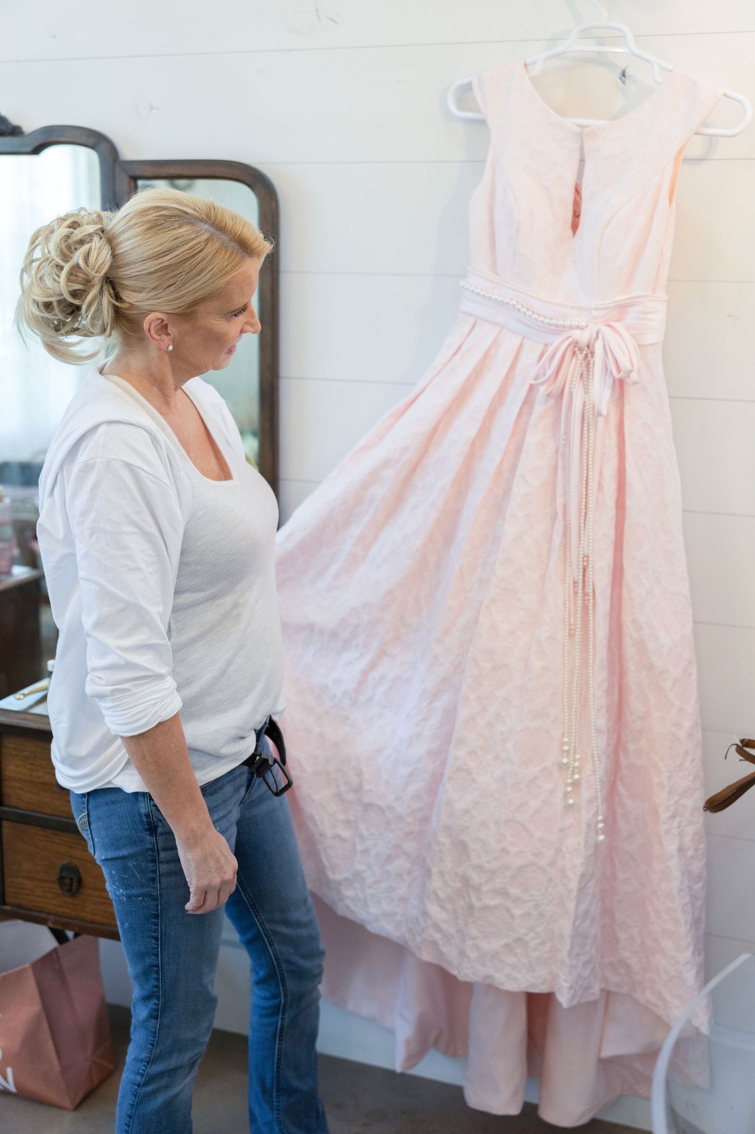 Blonde woman in white top and jeans admiring a pink formal gown hanging on a wall in a boutique.