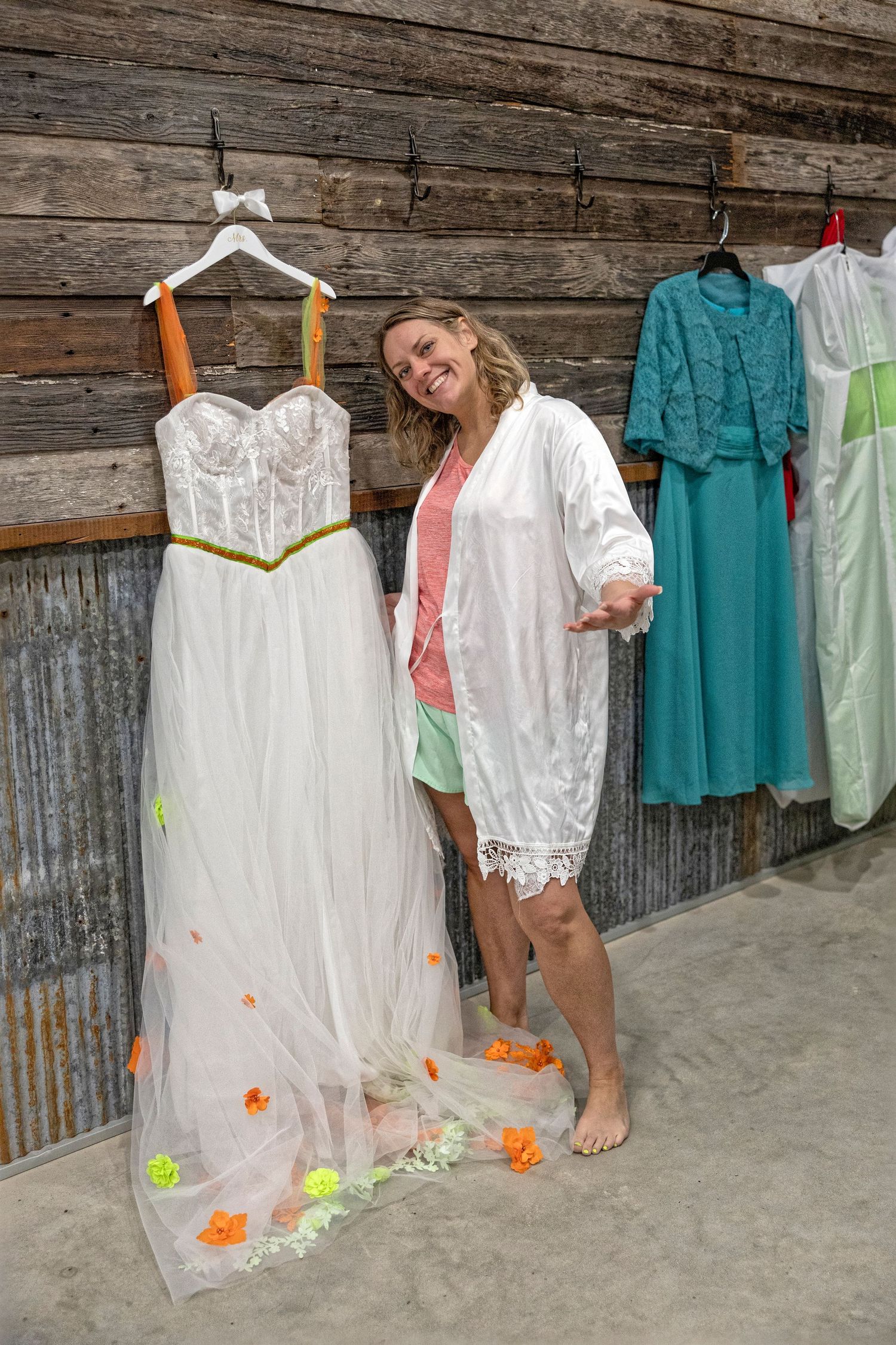 Smiling bride in white robe posing next to elegant wedding dress with orange accents hanging in rustic barn.