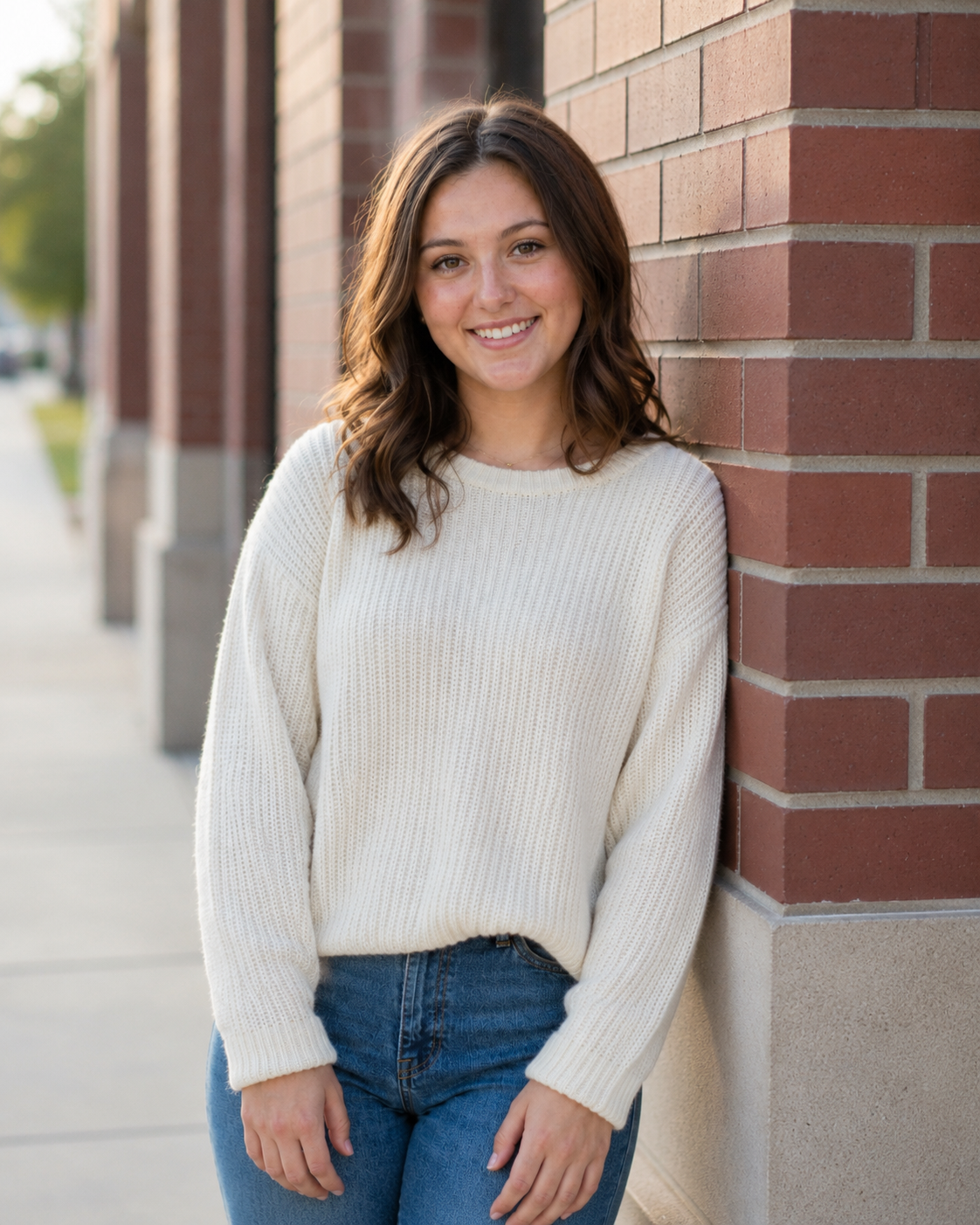 Young woman in cream knit sweater and jeans smiling against brick wall outdoors.