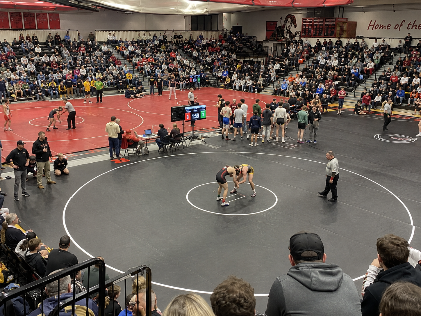 Wrestlers compete on center mat in packed high school gymnasium during a wrestling tournament.