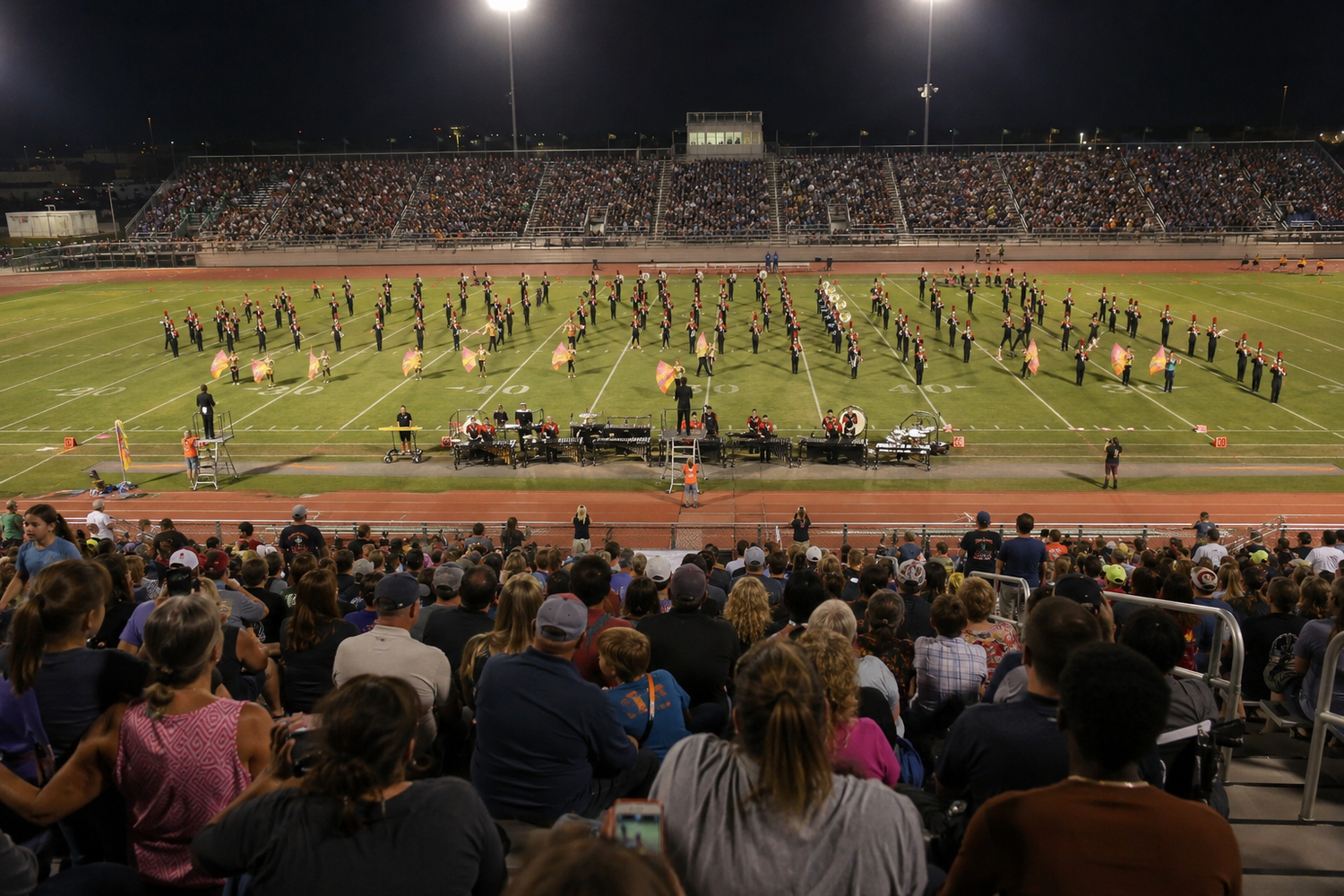 Large crowd watches a marching band perform on a football field during a nighttime stadium event.