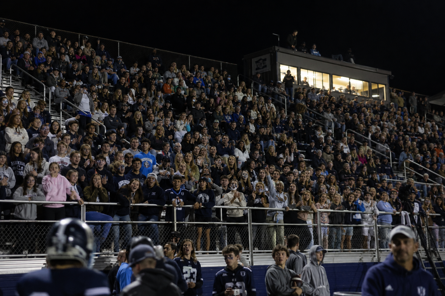 Large crowd fills bleachers at night football game under stadium lights.