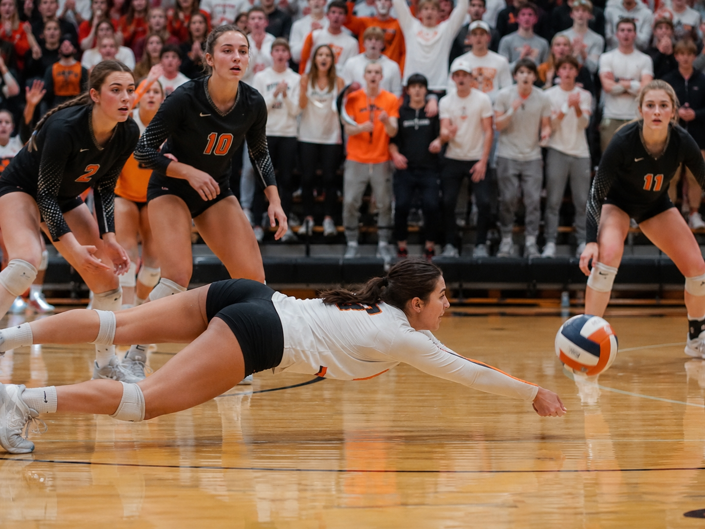 Volleyball player dives to make a dramatic dig save during an intense high school gymnasium match.