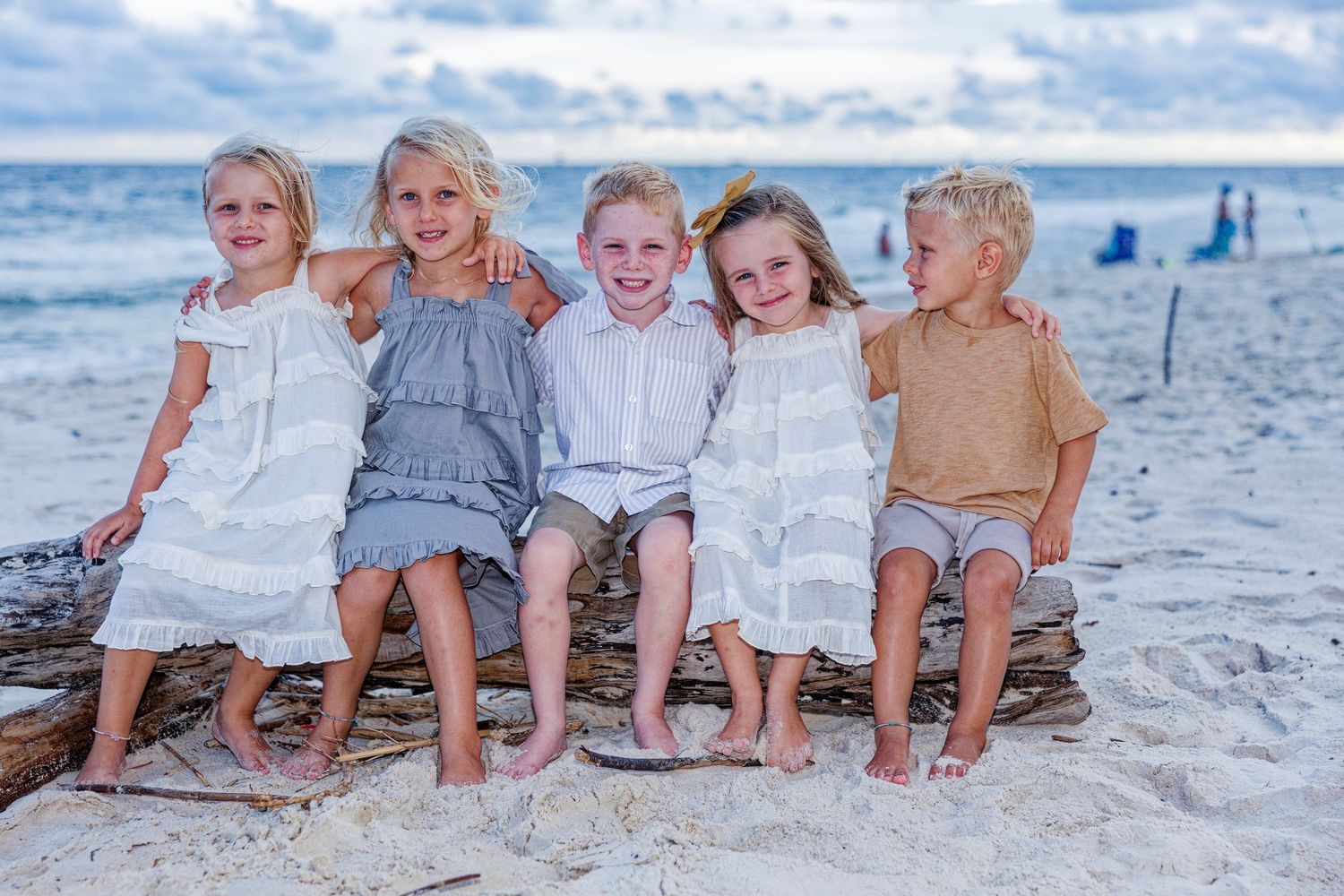 Children sitting together in a row on a sunny beach wearing coordinated white and blue outfits.