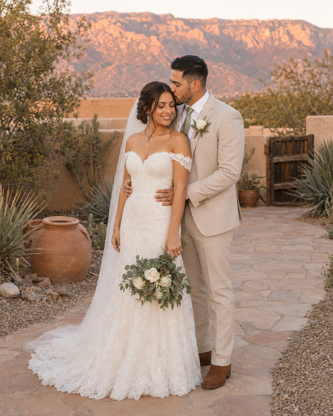 Groom in tan suit kisses bride in lace gown holding white bouquet at sunset desert wedding venue.