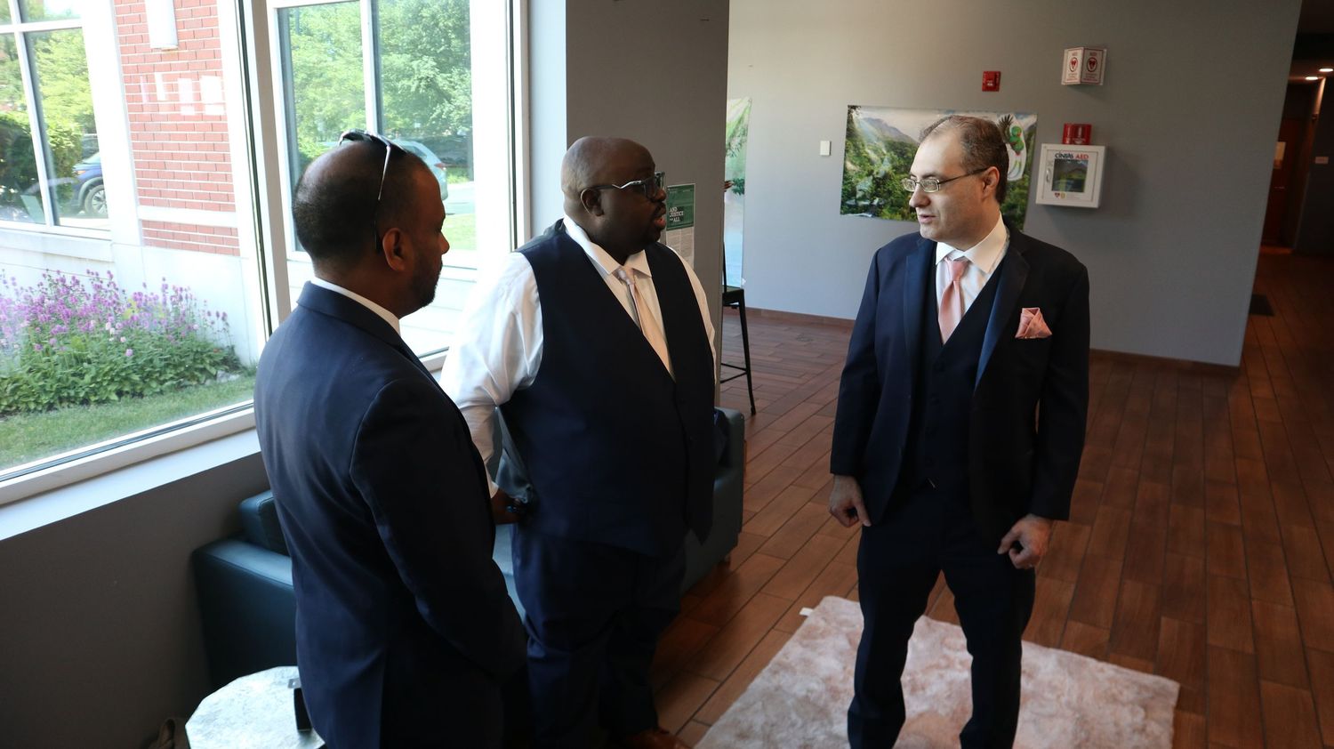 Three men in suits conversing indoors near a large window at a formal event or wedding.