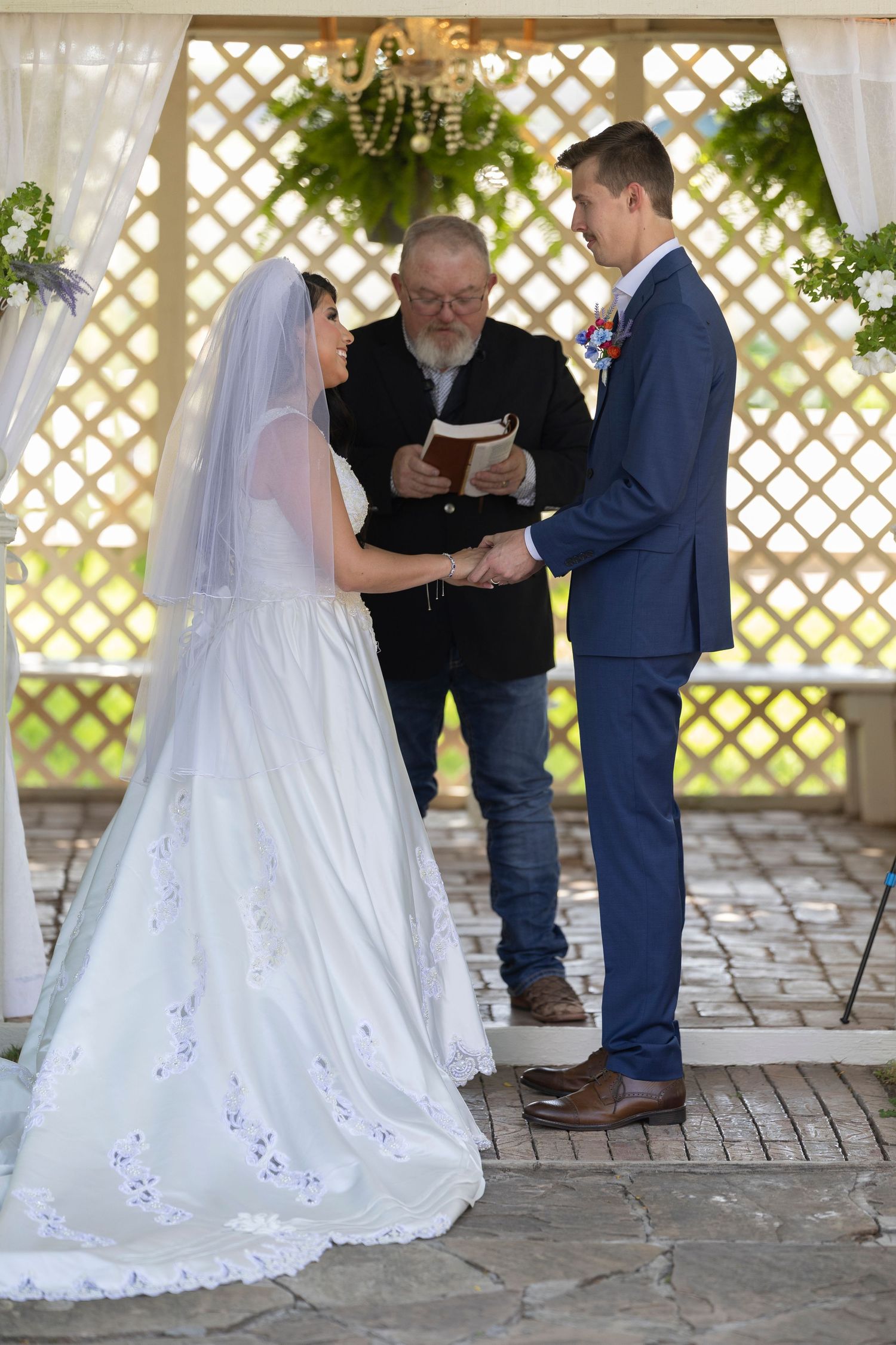 Bride and groom exchange vows at outdoor wedding ceremony with officiant reading from book under lattice arbor.