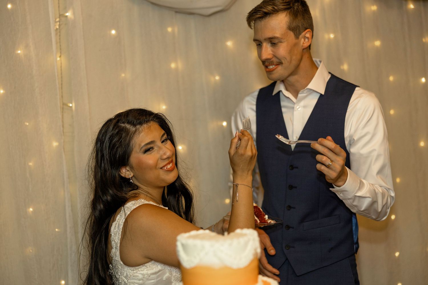 Couple cutting a wedding cake together, bride in white dress, groom in blue vest, fairy lights backdrop.
