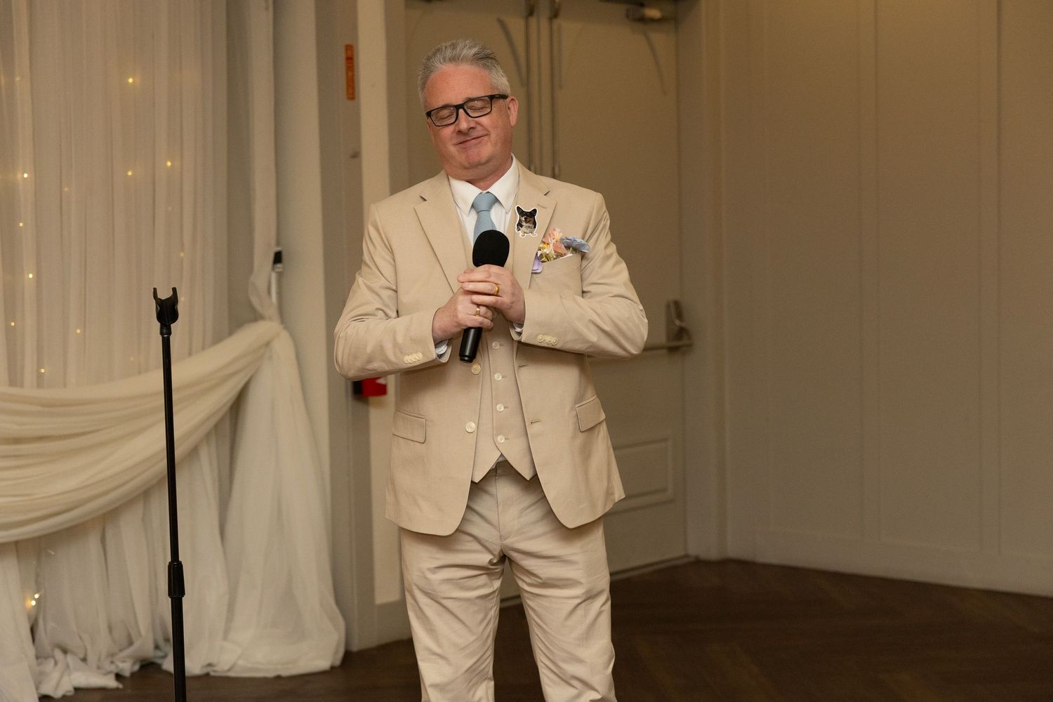 Man in beige suit speaking at wedding reception holding microphone on elegant white draped venue stage.
