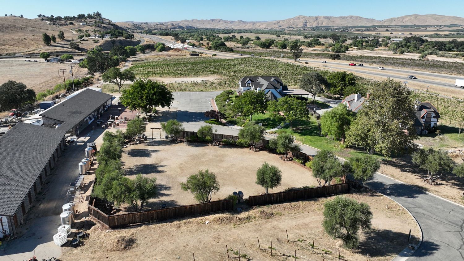Aerial view of a rural ranch property with vineyards, trees, and open land near a highway in a dry California landscape.