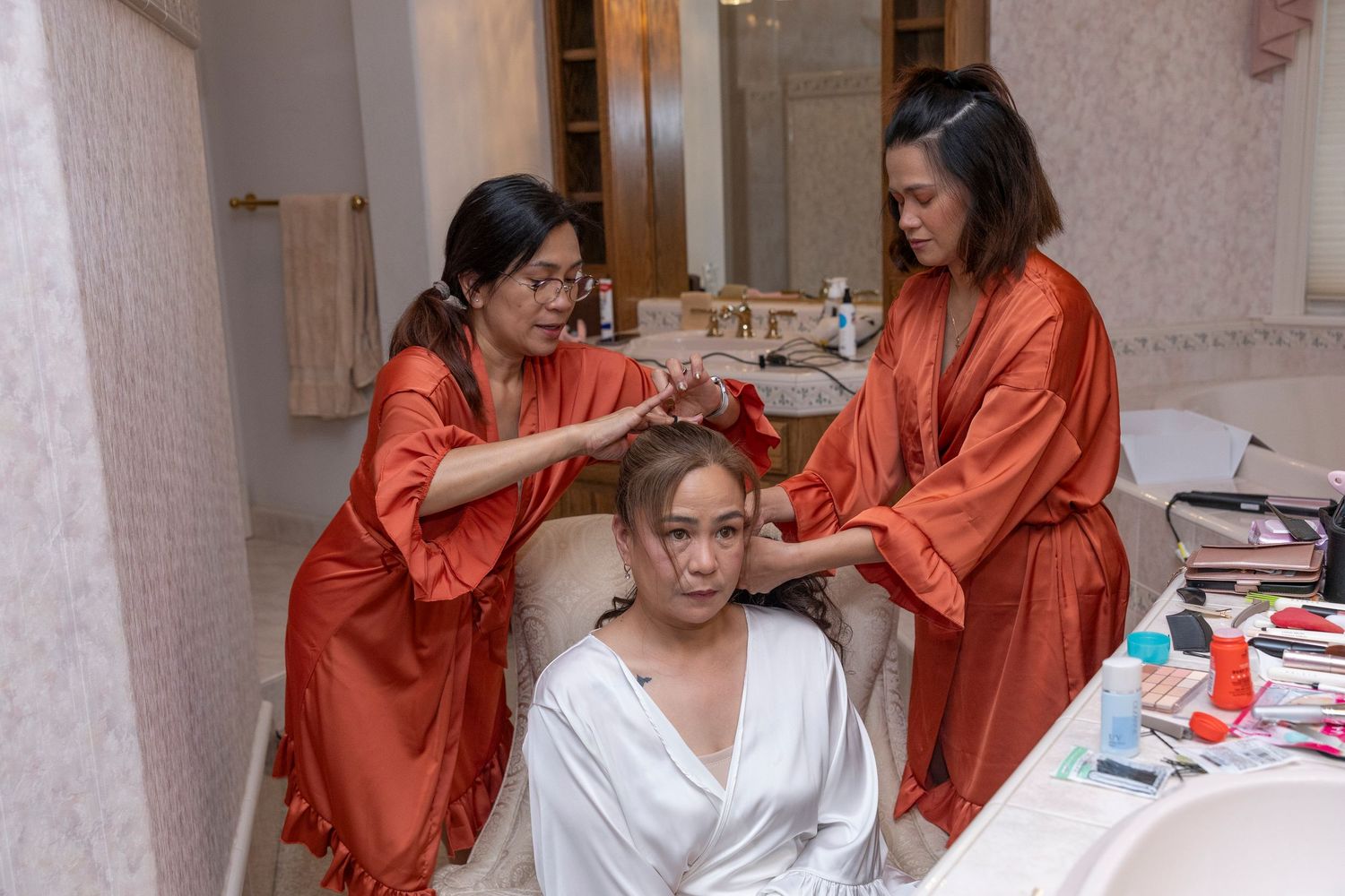 Two bridesmaids in orange robes styling a bride's hair and makeup before a wedding ceremony.