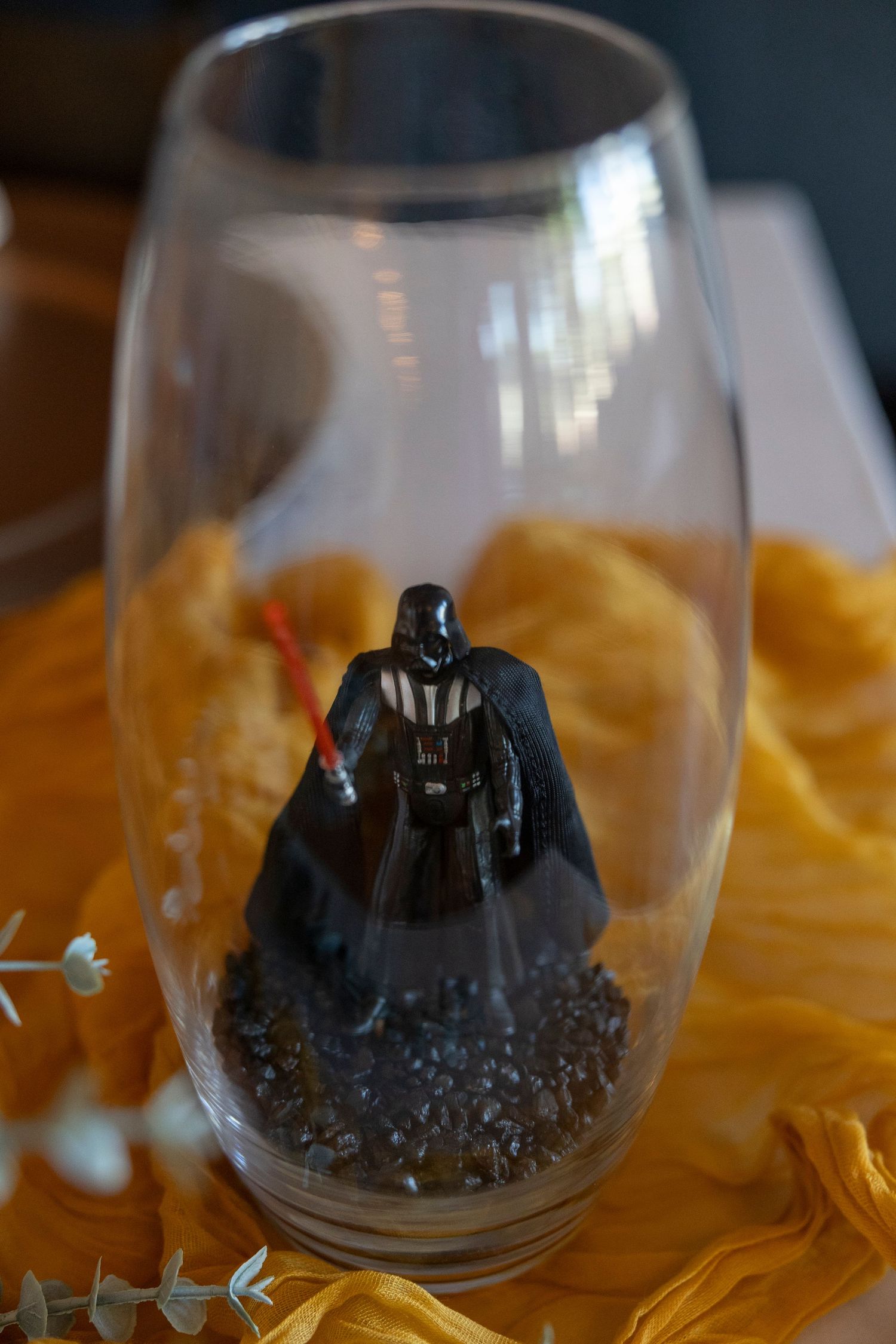 Darth Vader figurine inside a clear glass vase with gravel, surrounded by yellow fabric and eucalyptus leaves.