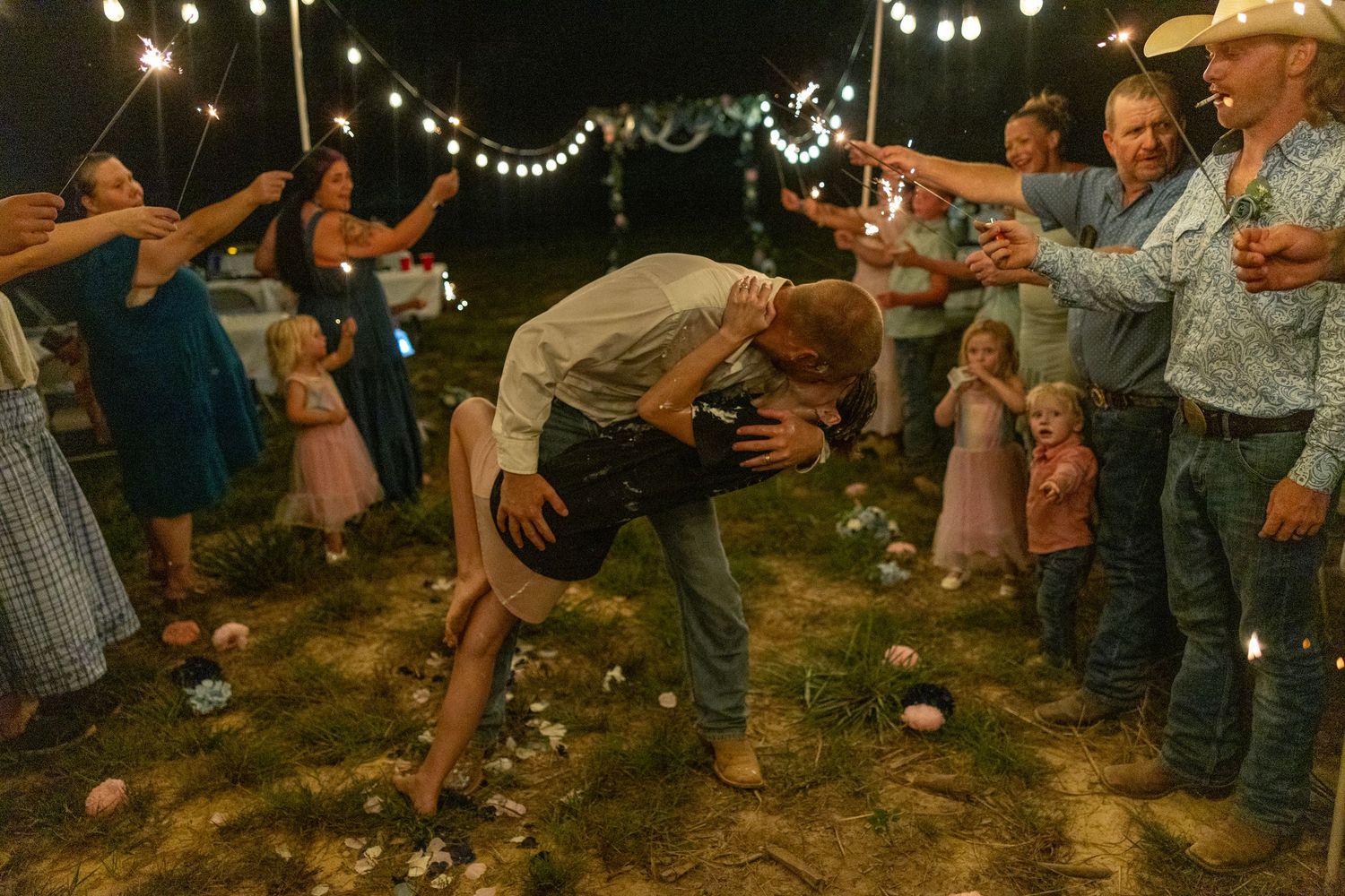 Couple shares romantic dip kiss at outdoor wedding reception with sparklers and guests cheering under string lights at night.