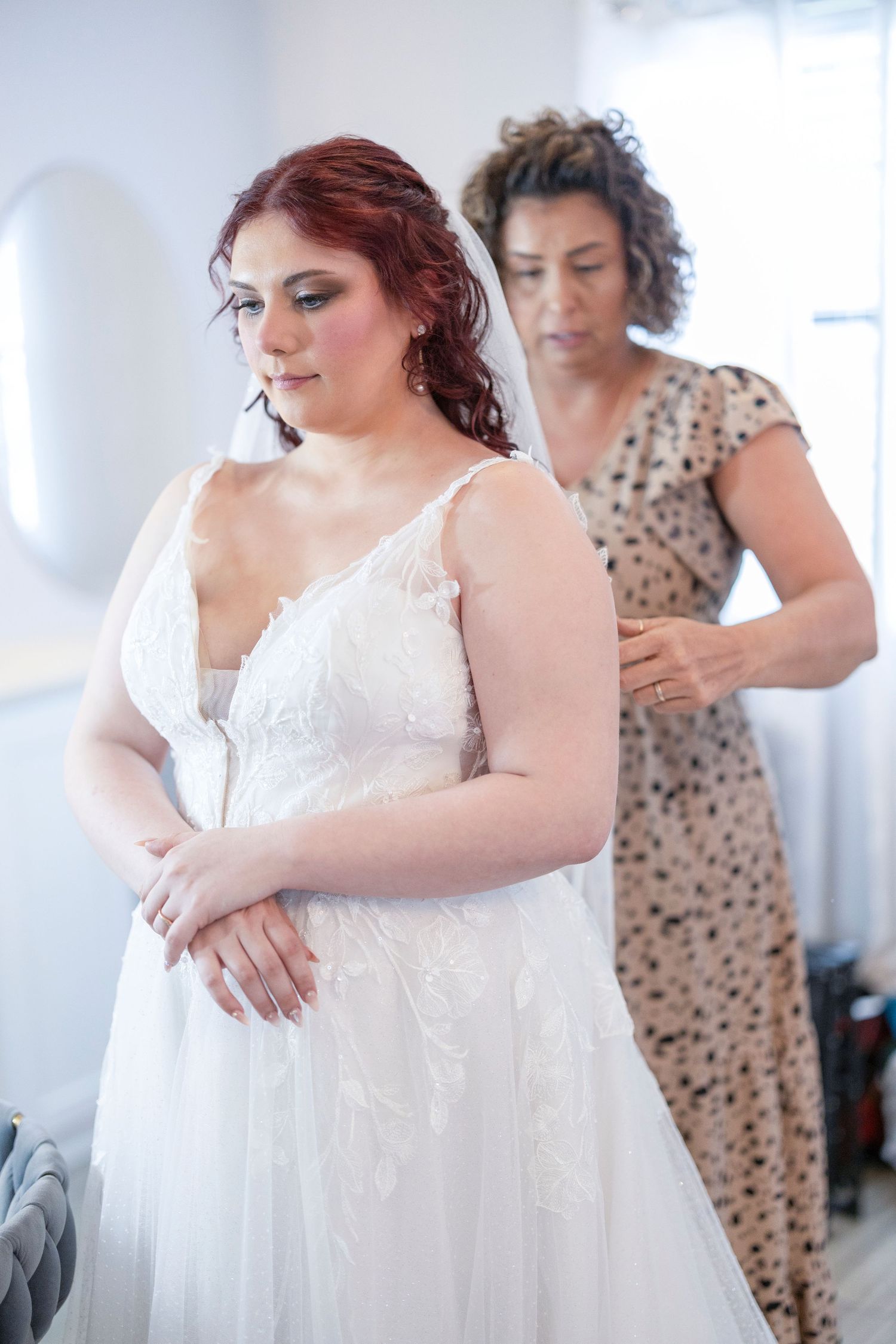 A bride in a white lace wedding dress gets help with final adjustments during wedding preparations.