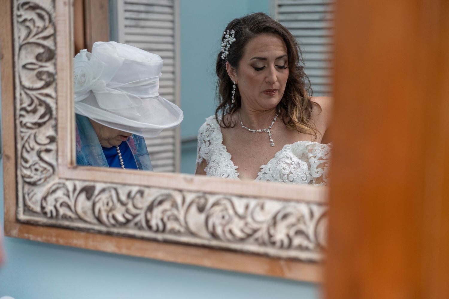 A person in a white lace wedding dress adjusts their hair in front of an ornate mirror.