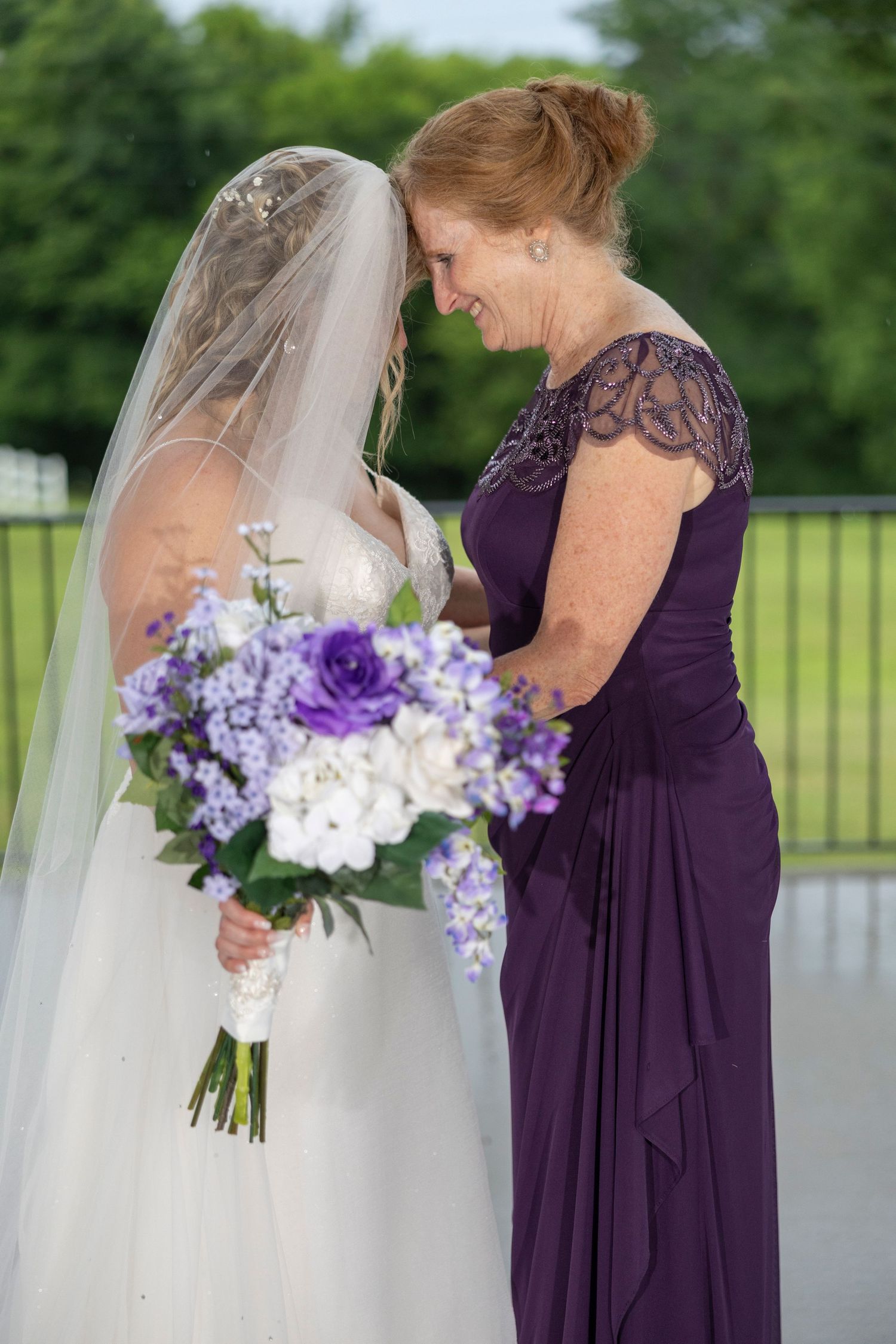 Wedding party members in deep purple bridesmaid dresses holding purple and white floral bouquets outdoors.