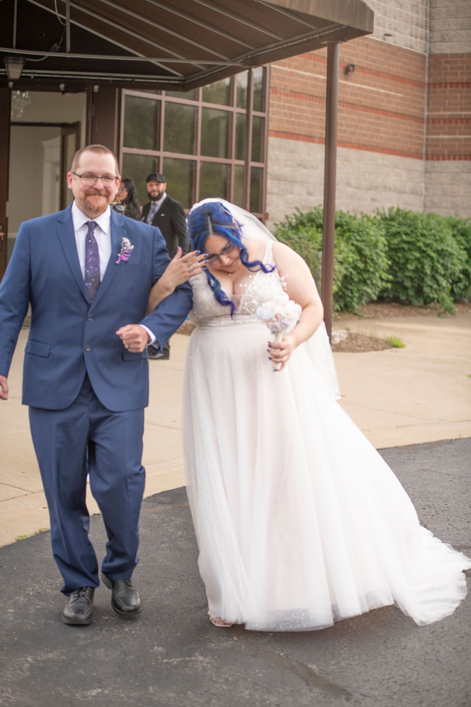 A wedding couple walks together outside a building entrance, with the bride wearing a white gown and having blue hair.