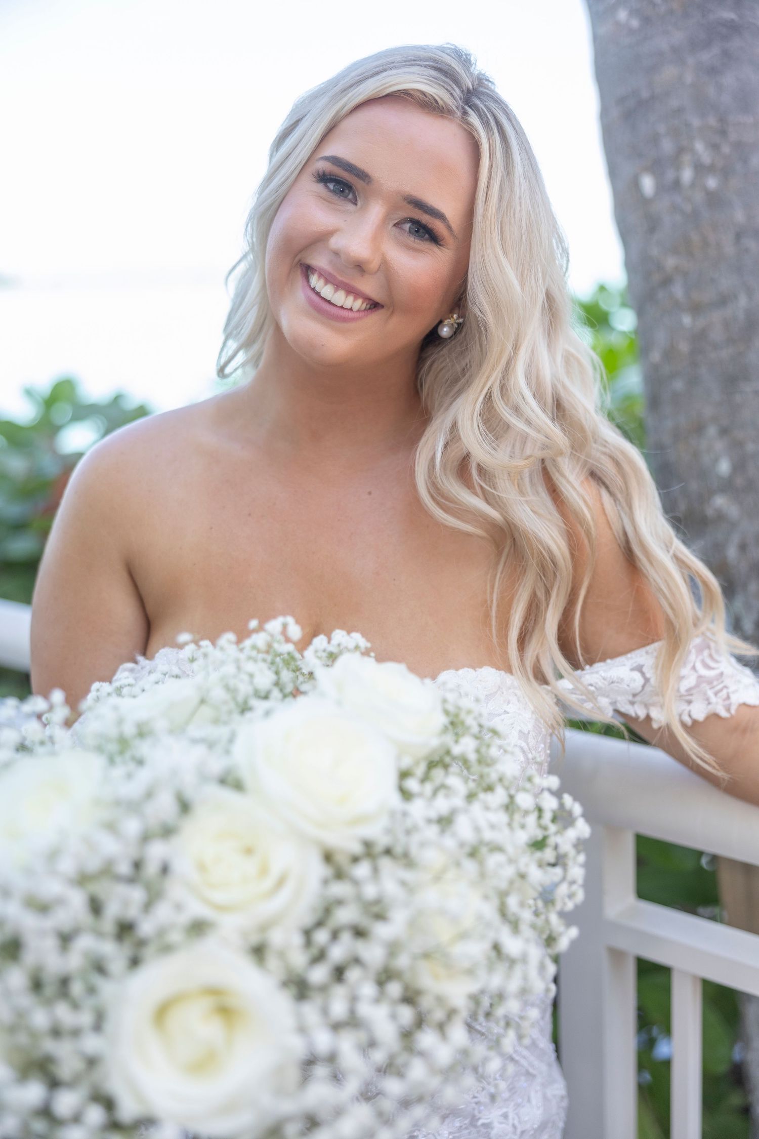 A bride in a strapless lace wedding dress holds a white rose and baby's breath bouquet while smiling outdoors.