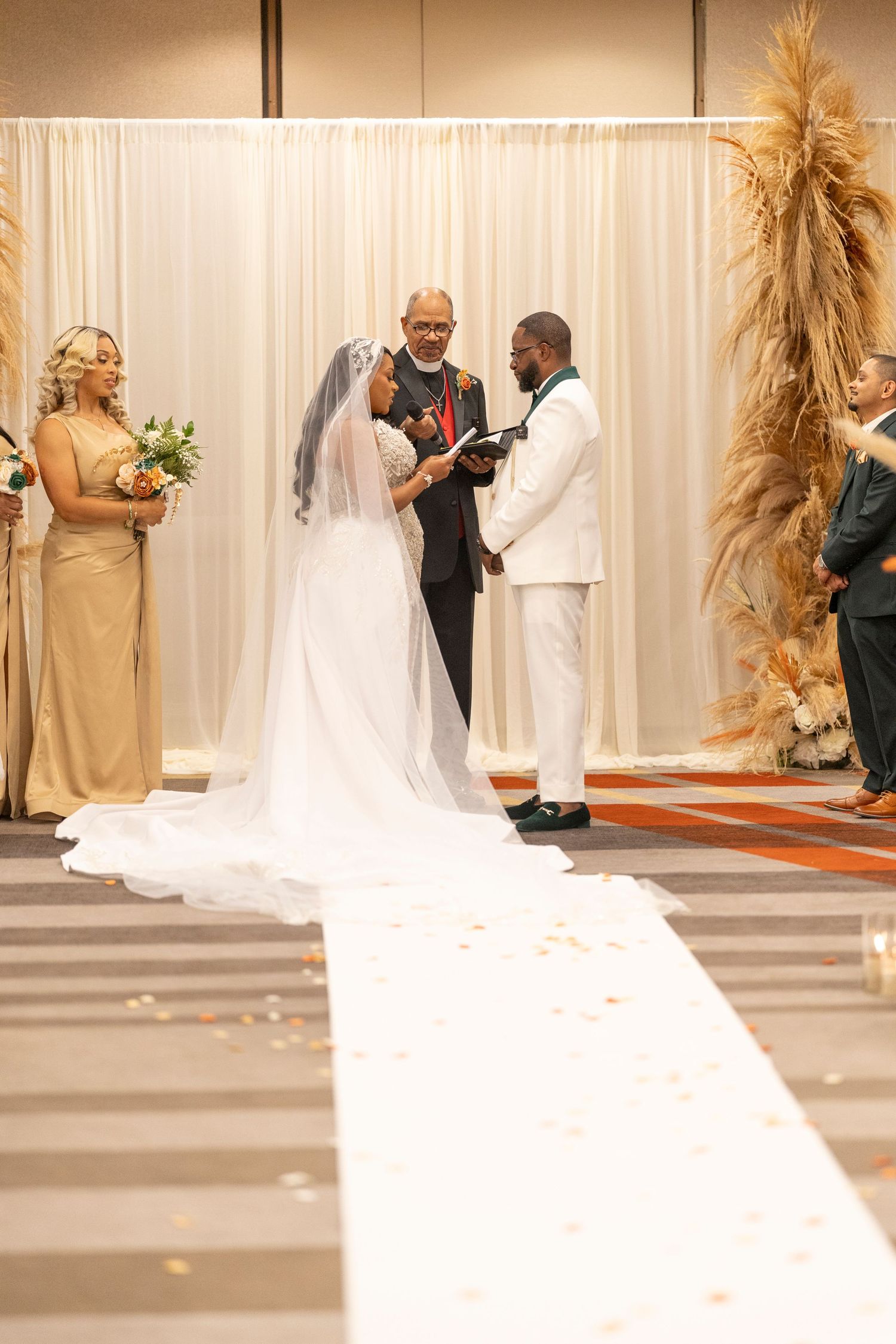 A wedding ceremony takes place in front of a white backdrop with pampas grass decor and a long flowing bridal train.