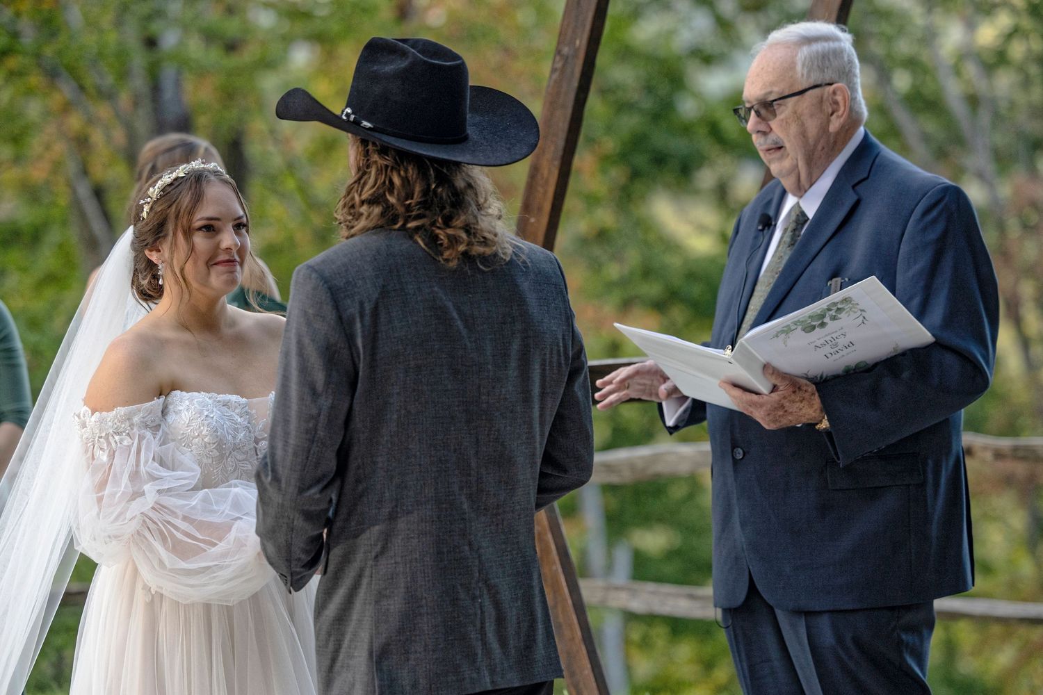 An outdoor wedding ceremony takes place in a natural setting with a bride in white dress and officiant holding a book.