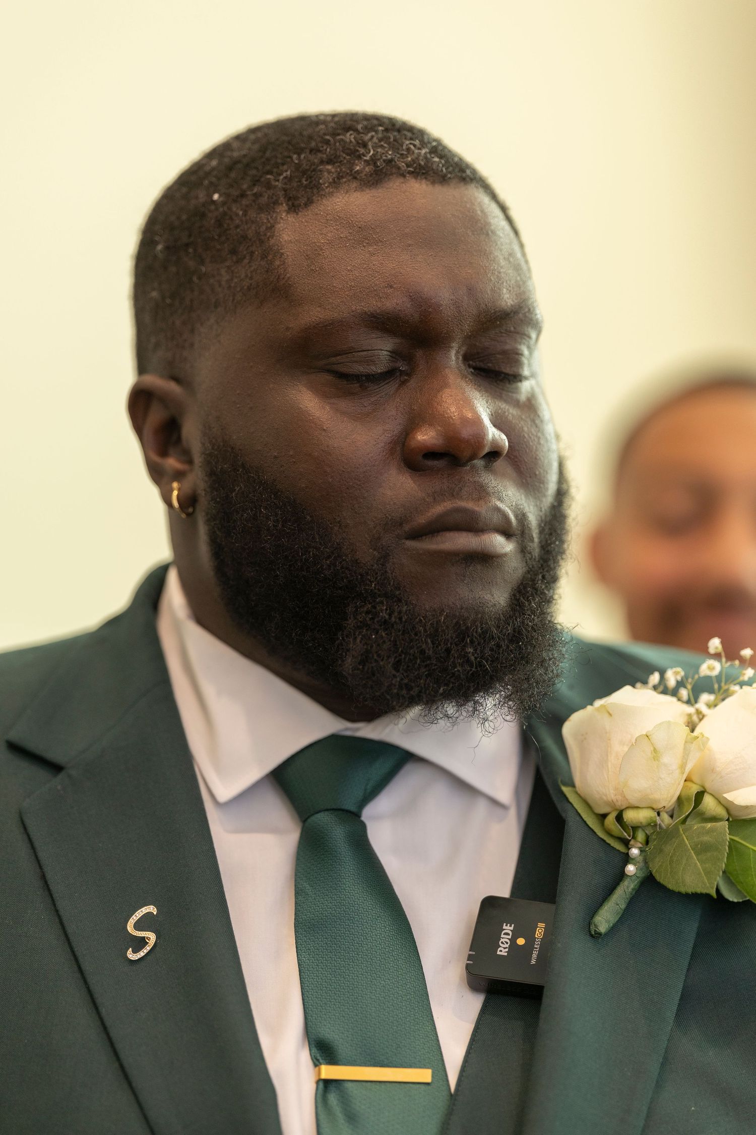 A person in a dark green suit with a matching tie and white boutonniere closes their eyes during an emotional moment.