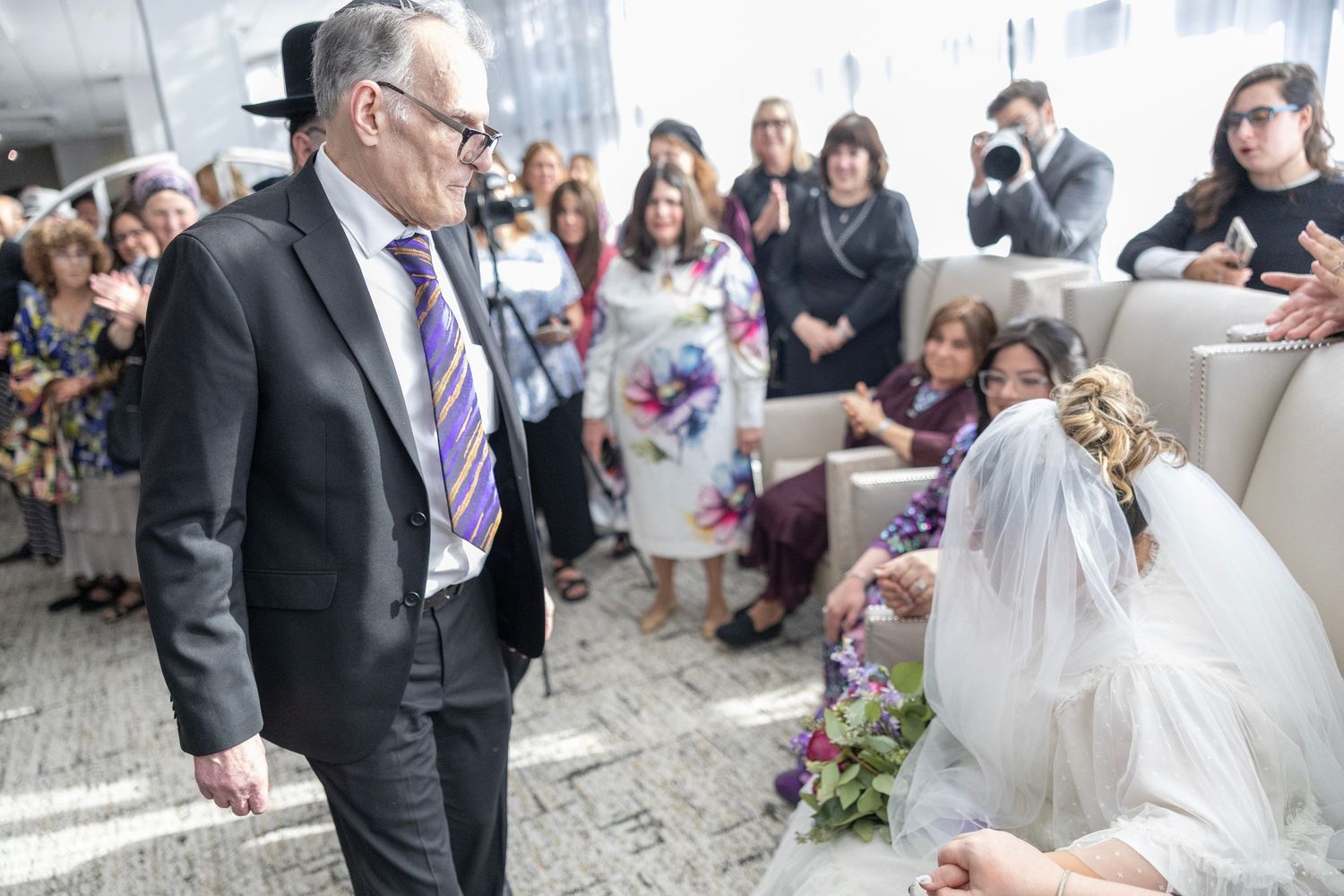 Wedding guests gather around to watch a couple sign their marriage certificate during an indoor ceremony.