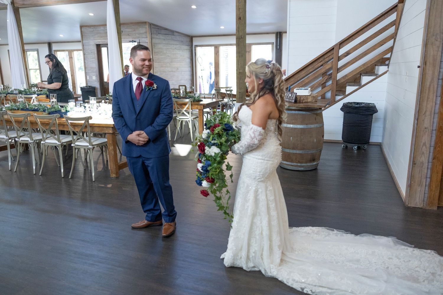 A couple shares their first look moment in a rustic barn venue with wooden barrels and gold chiavari chairs.
