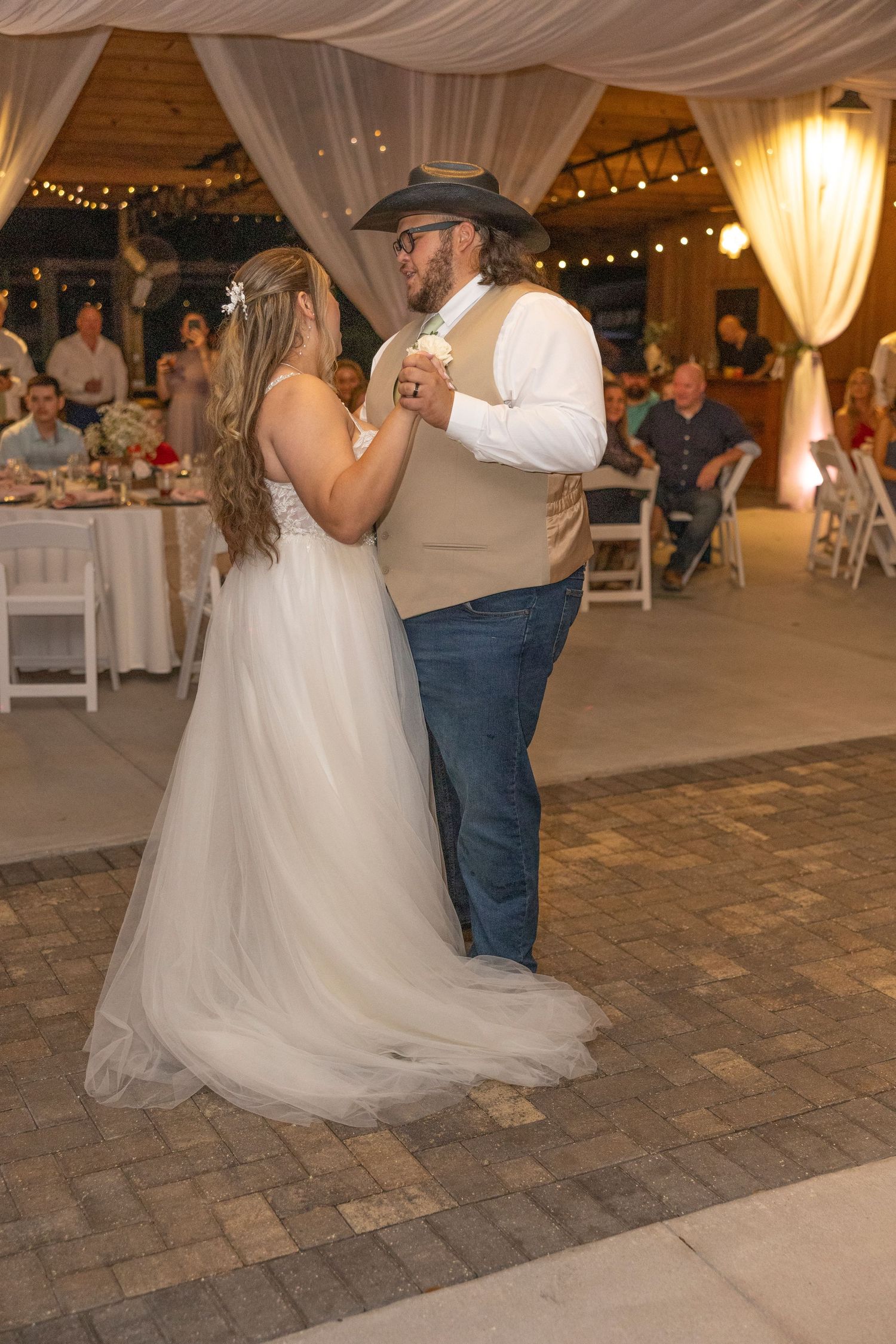 A couple shares their first dance at a rustic barn wedding reception with twinkling lights and white drapes.