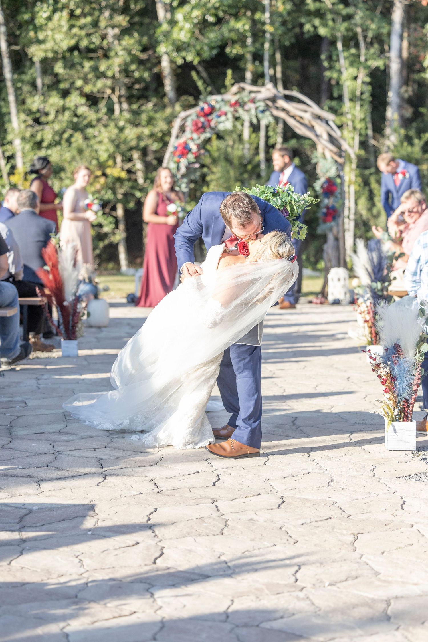 Wedding couple dips dramatically during outdoor ceremony in wooded setting with guests watching from both sides.