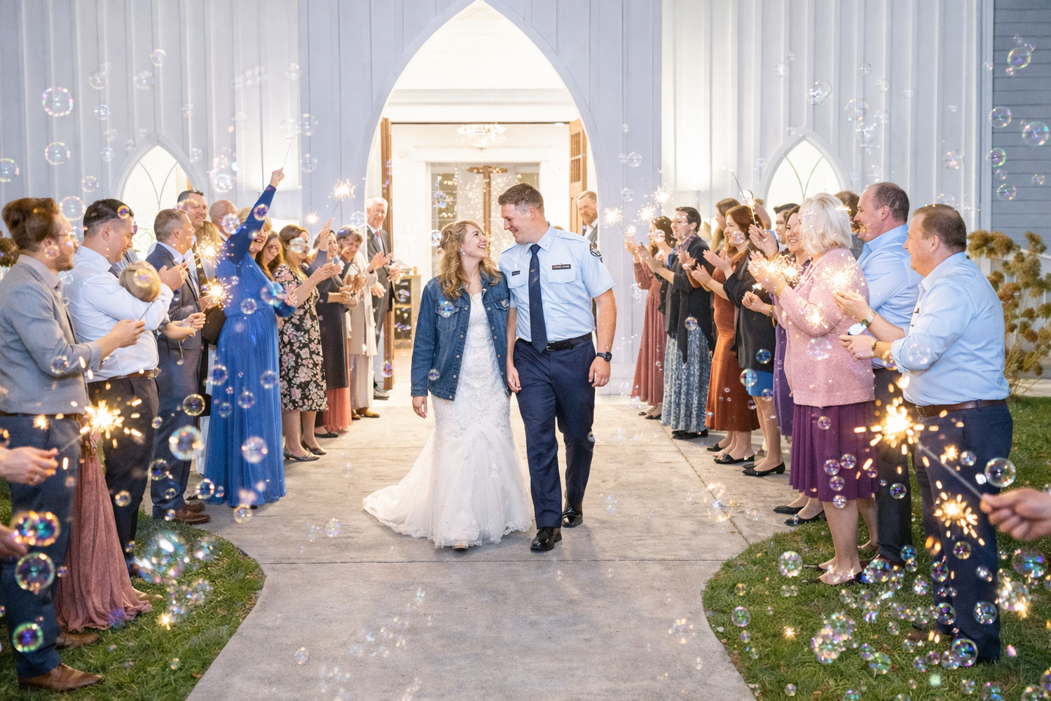 Wedding guests hold sparklers forming an aisle as newlyweds exit a white chapel during their celebration.